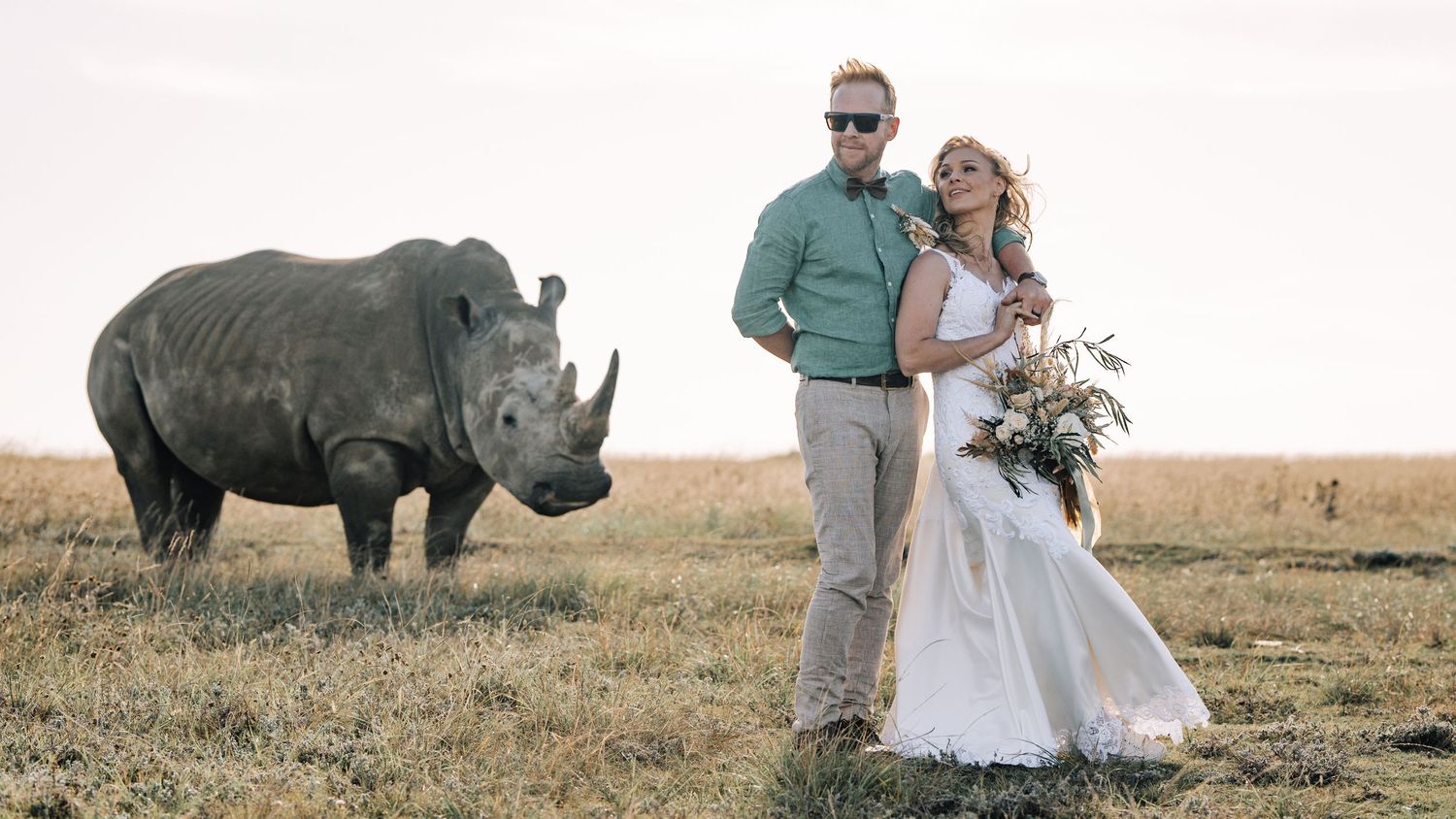 Wedding couple poses in a grassy field with a white rhinoceros standing nearby in the background.