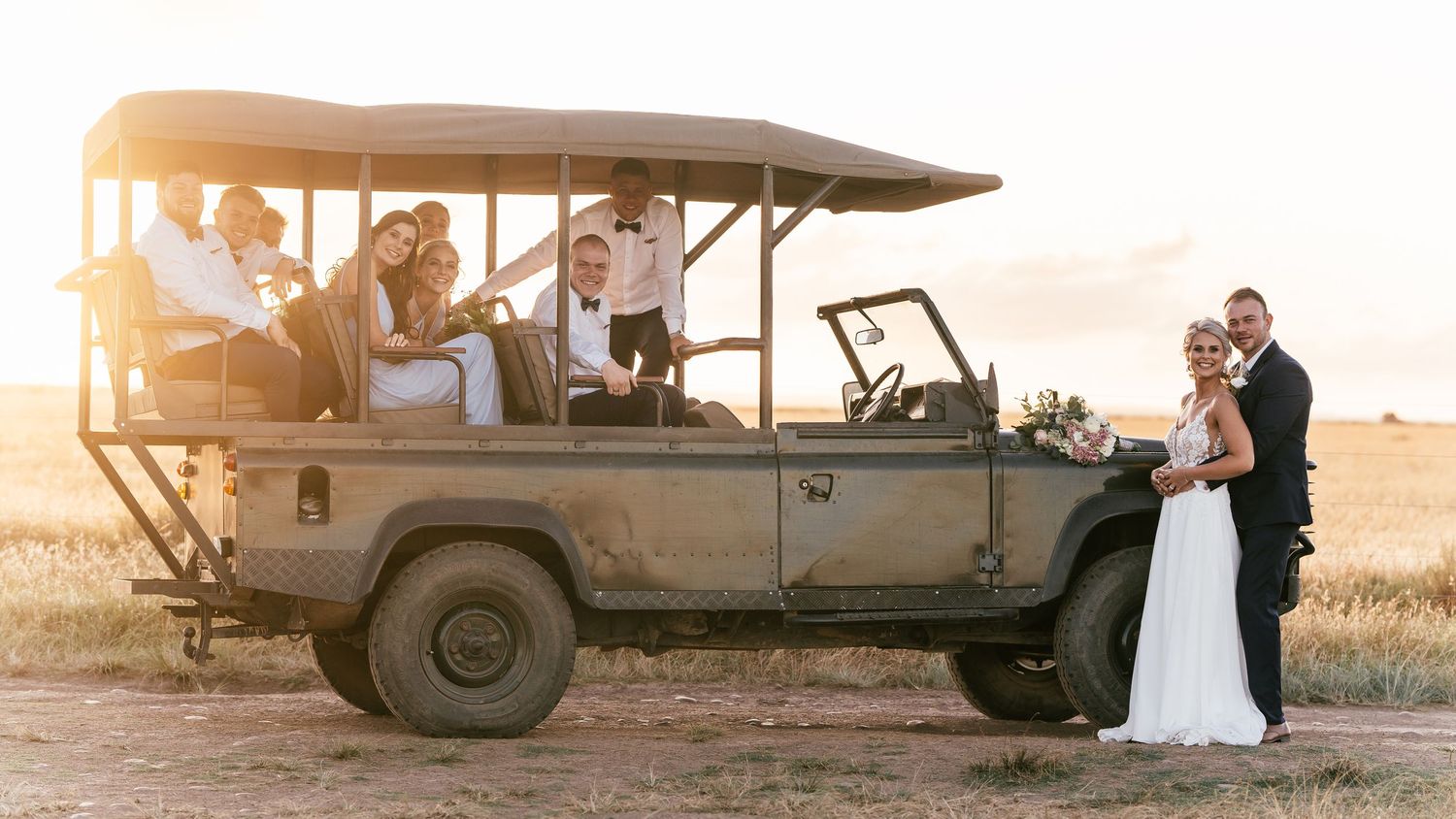Wedding party posing on a safari jeep at golden hour in an open field.