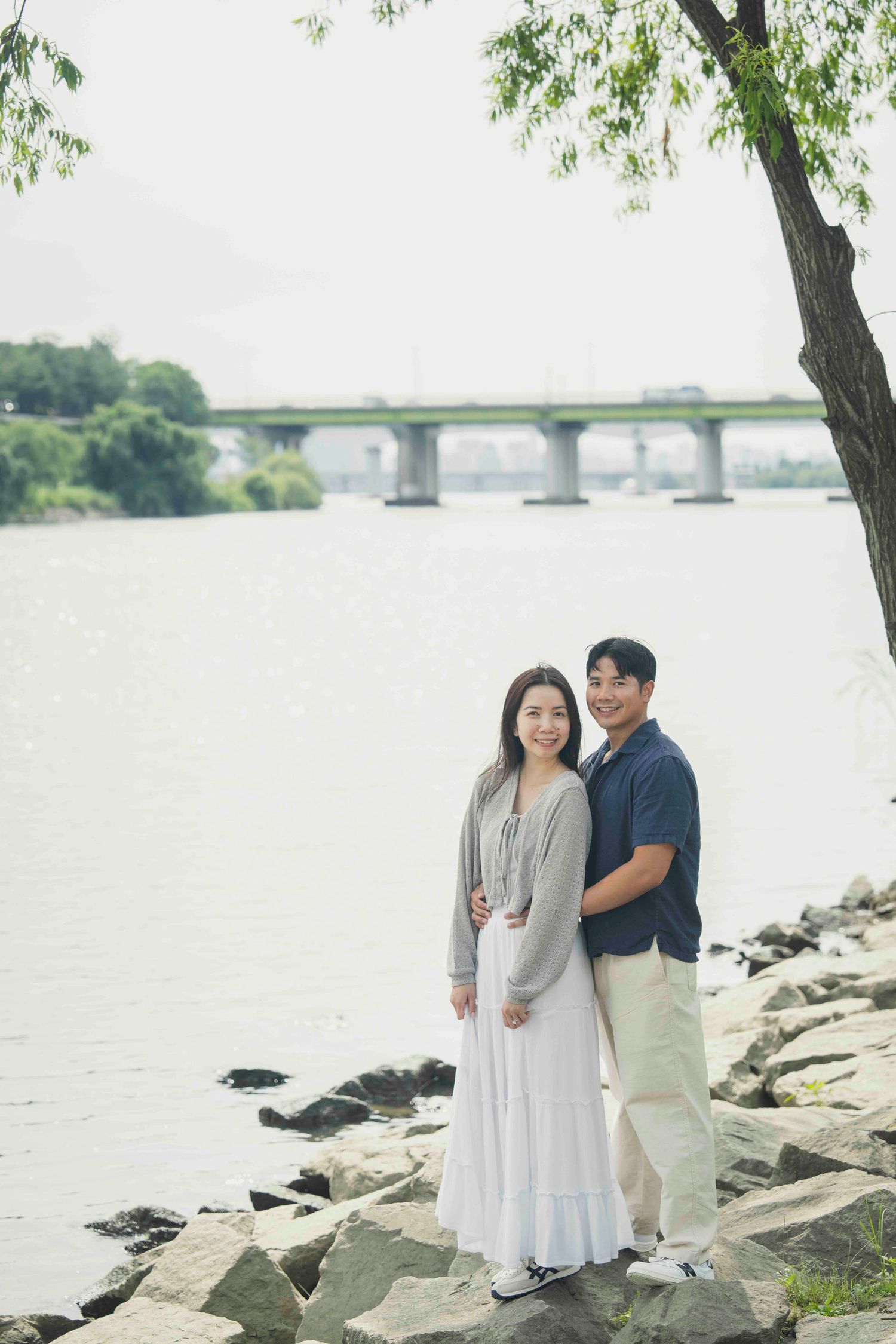 Asian couple standing on rocky riverbank near bridge, smiling at camera on overcast day.