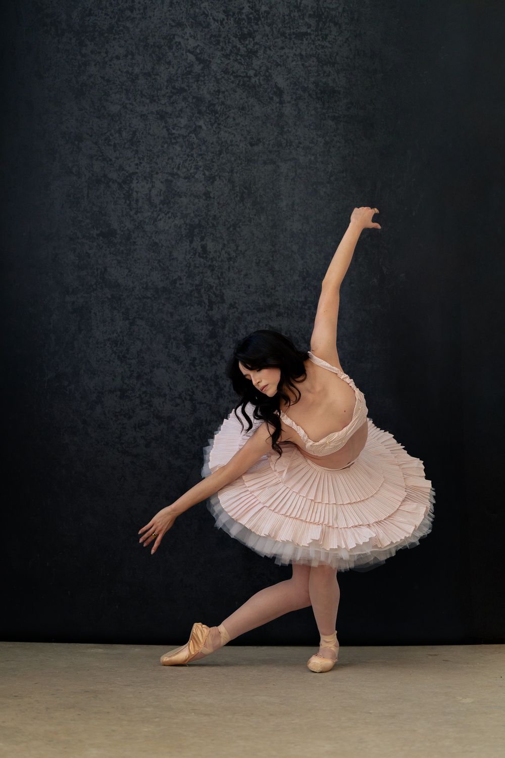 A ballerina in a white tutu performs an elegant arabesque pose against a dark textured studio backdrop.