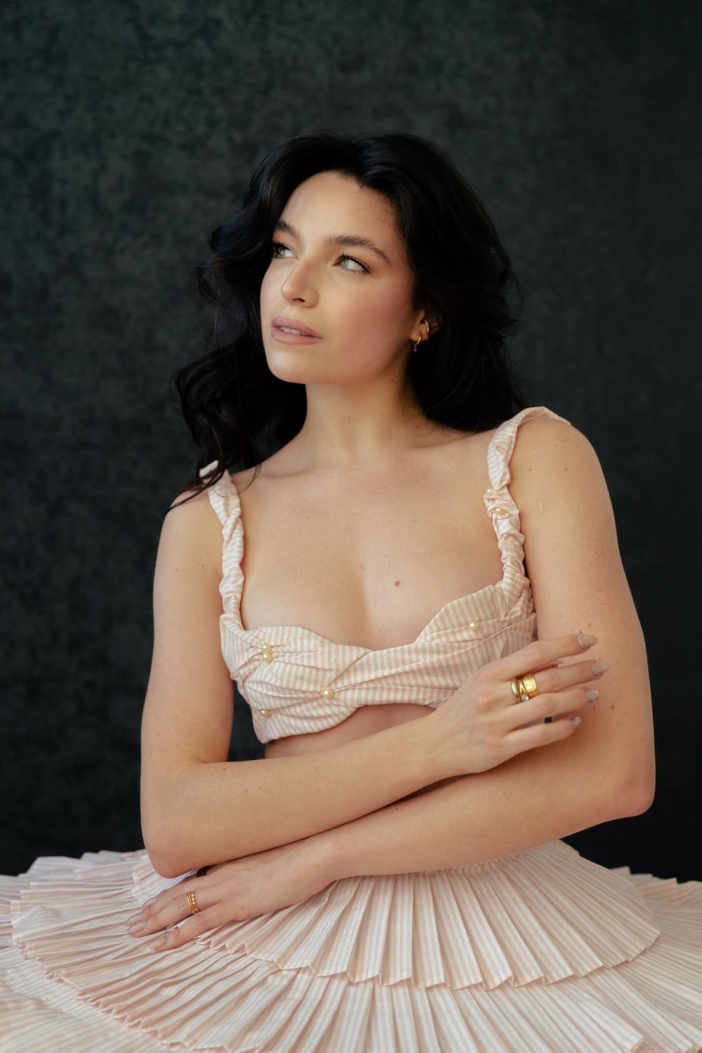 A dark-haired woman in a cream ruffled corset top poses thoughtfully with crossed arms against a dark green background.