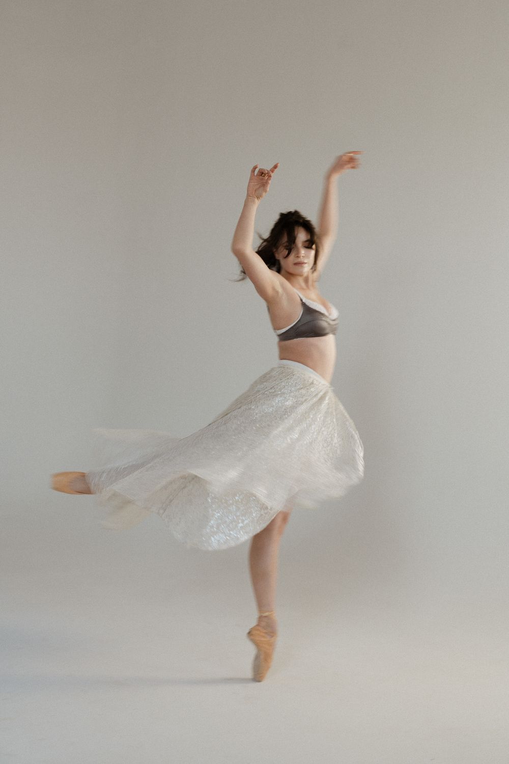 A graceful ballerina in a gray bra top and white lace skirt performs an elegant en pointe pose with arms raised.