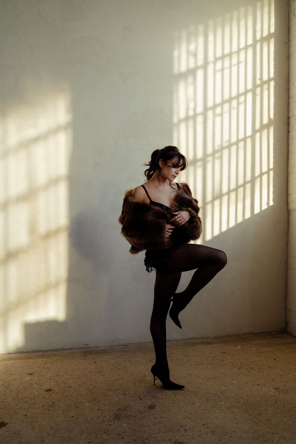 Woman in black lingerie and fur coat poses dramatically in sunlit warehouse with high heels.