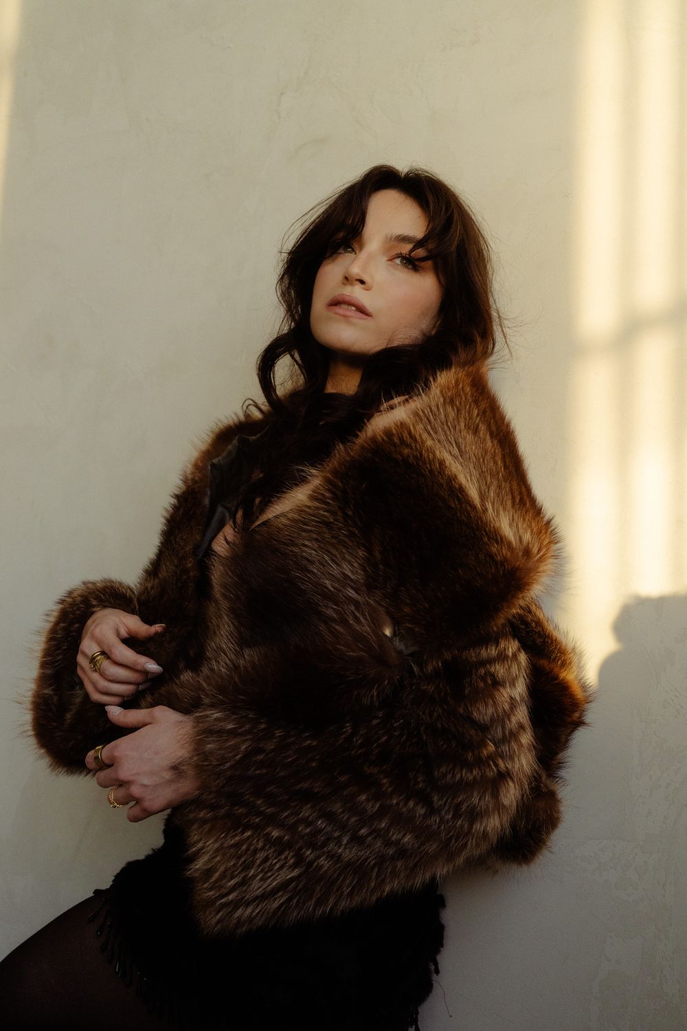 A brunette woman in a luxurious brown fur coat poses confidently against a warm sunlit white wall.