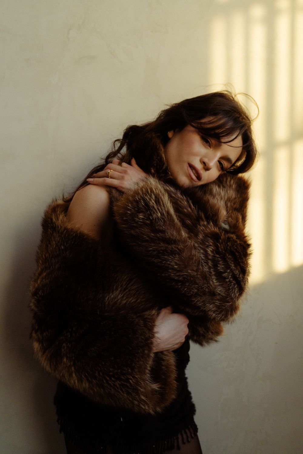 A woman in a brown fur coat leans against a wall, eyes closed, bathed in warm sunlight.