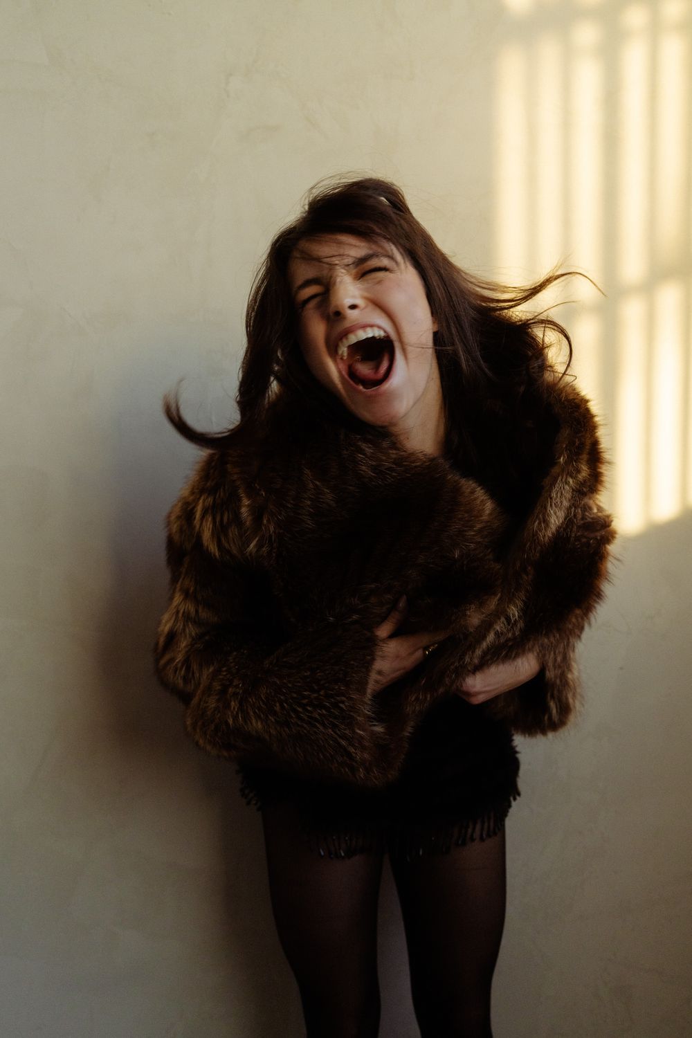 Woman in brown fur coat laughing with eyes closed, hair flowing, against a sunlit wall.