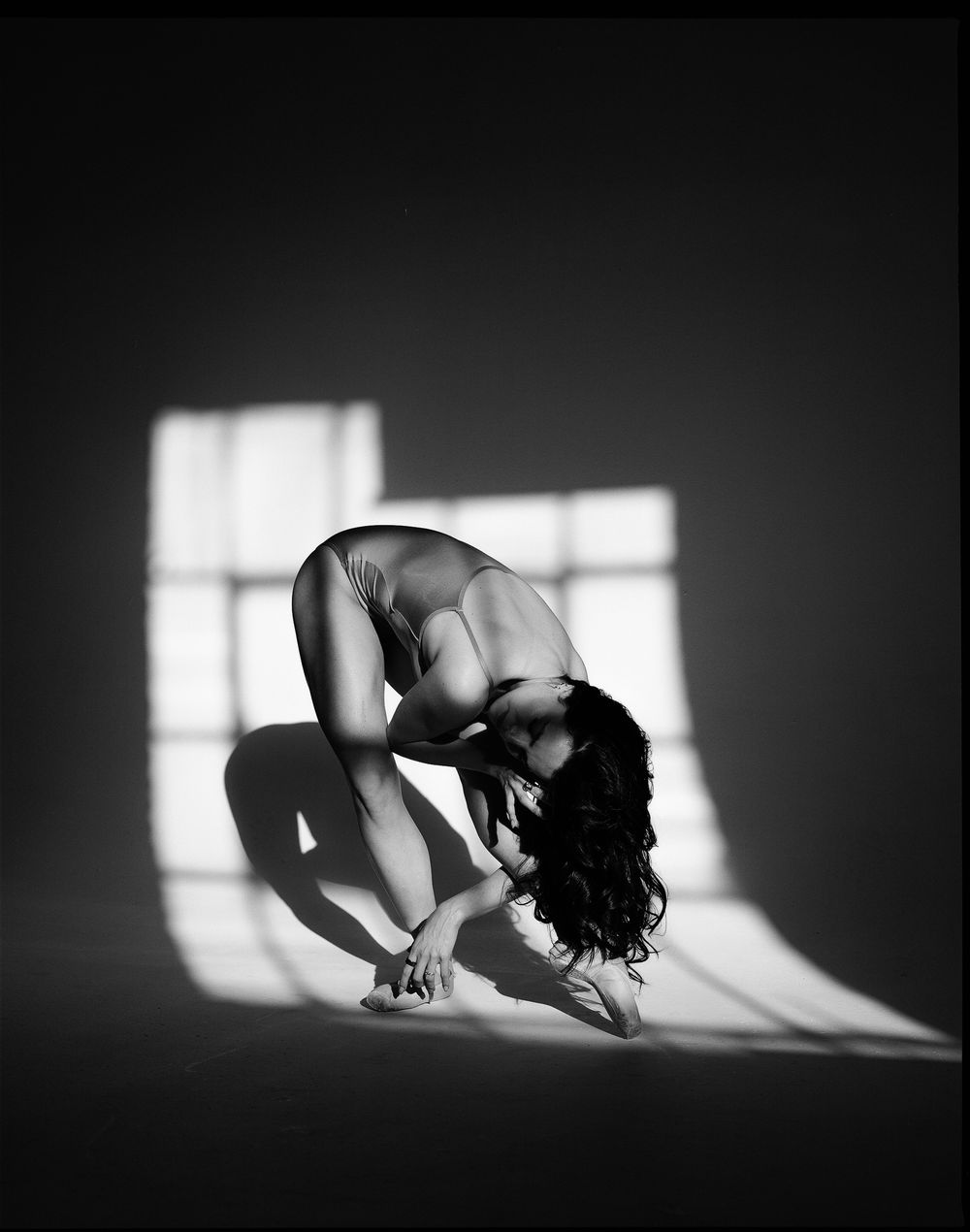 Black and white photo of a nude figure gracefully bent forward, illuminated by dramatic window light casting geometric shadows.