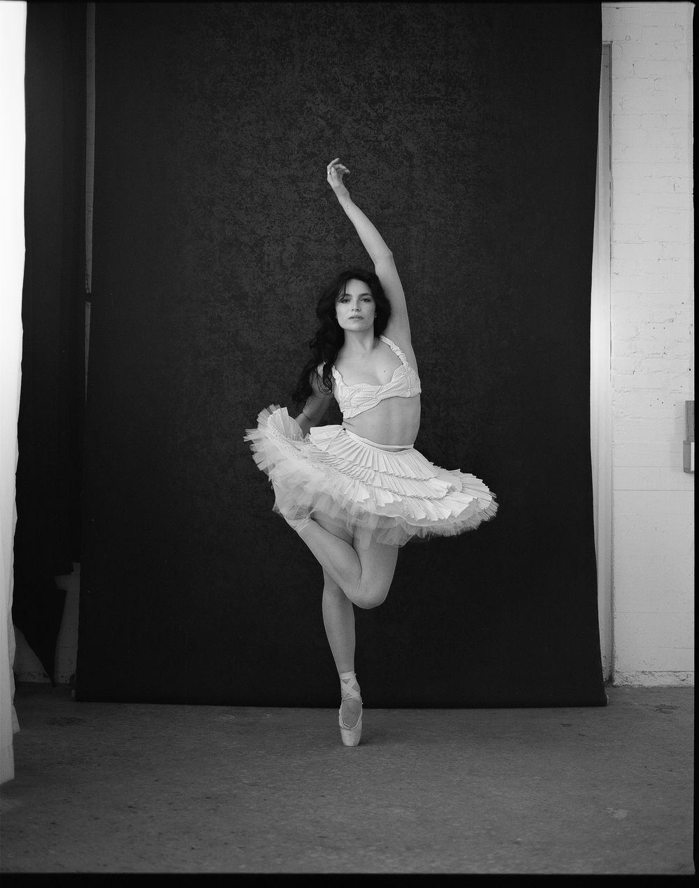 A ballerina en pointe in a white tutu and crop top performs an arabesque against a dark studio backdrop.