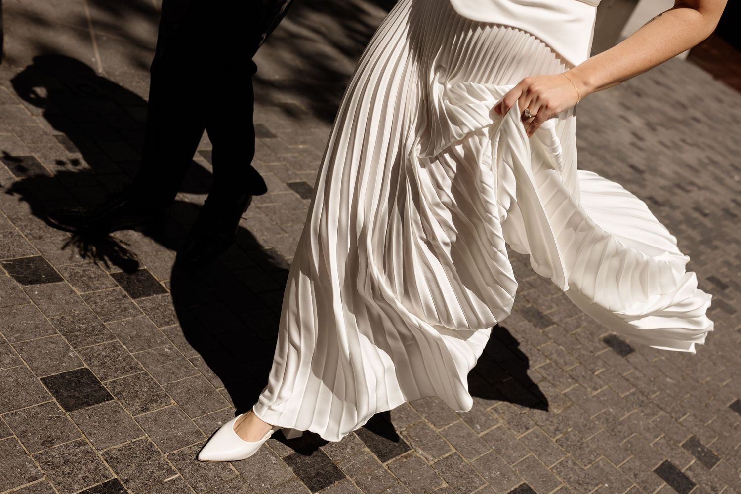 Woman in white pleated midi skirt and white heels walking on cobblestone pavement in bright sunlight.