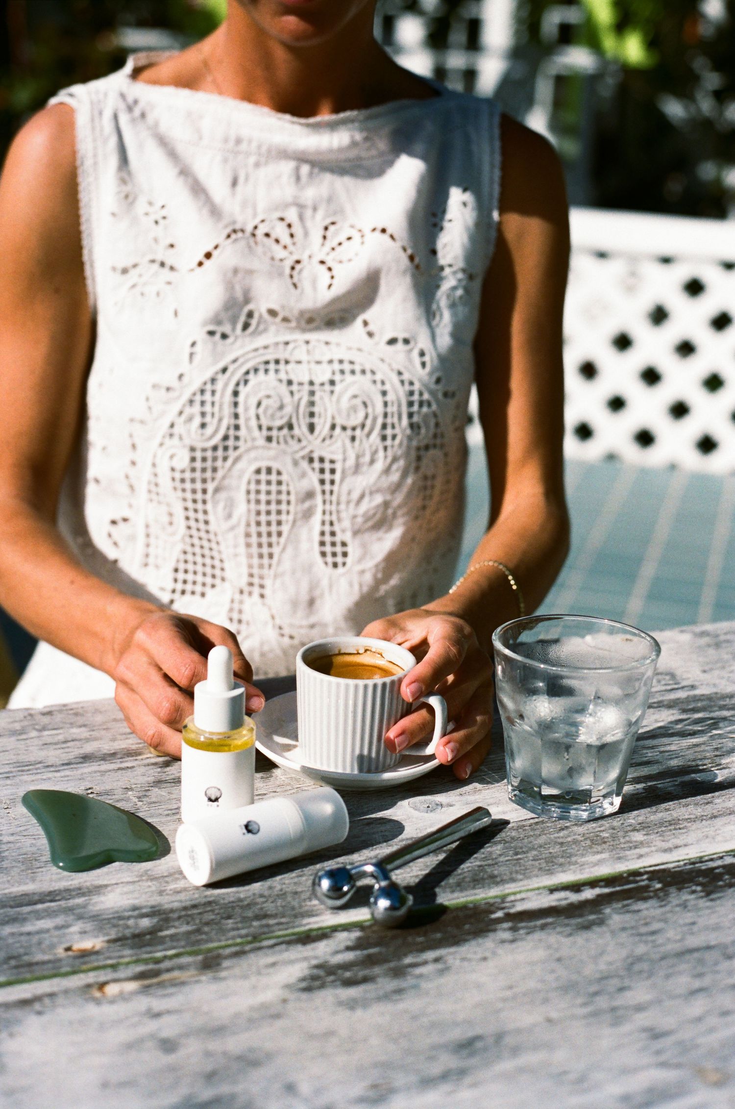 Woman in white lace top holding espresso cup outdoors with skincare products and iced water on rustic table.