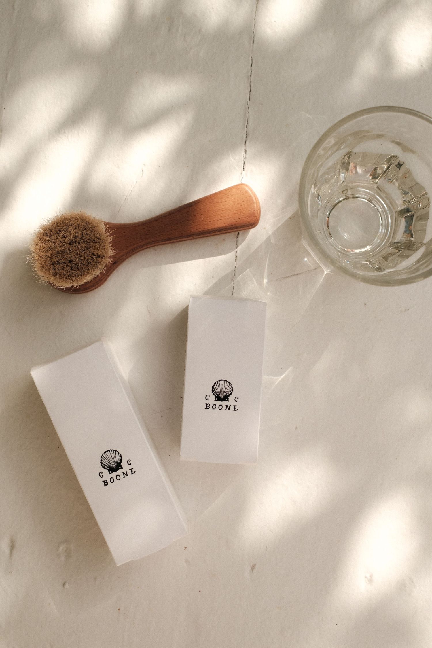 Two white Coco Boonz product boxes beside a wooden facial brush and glass on a sunlit white surface.