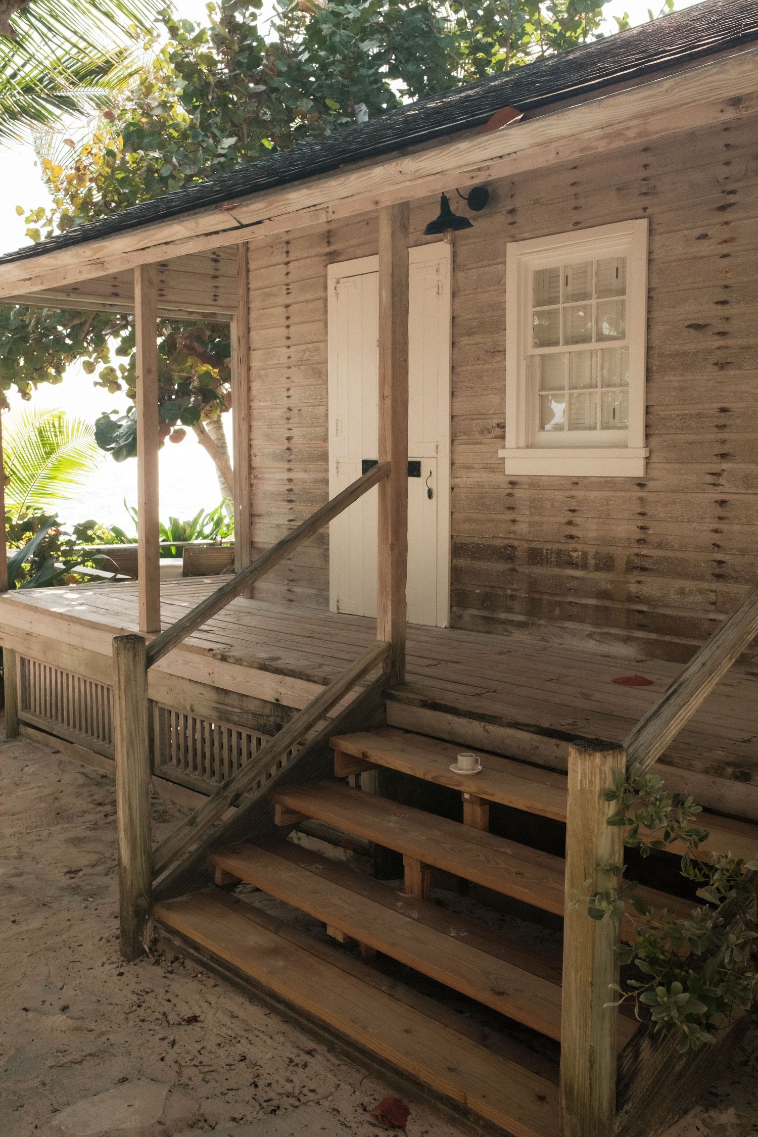 Rustic wooden beach cottage with weathered siding, covered porch, white door, and sandy steps surrounded by tropical trees.