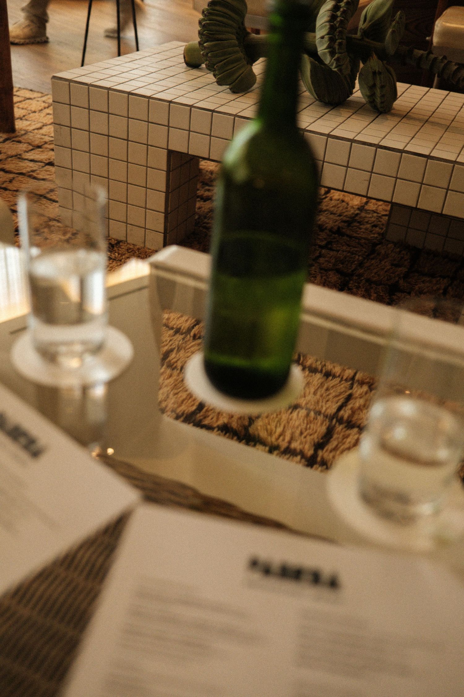 A green glass bottle and water glasses on a restaurant table with menus, tiled bench and cactus in background.
