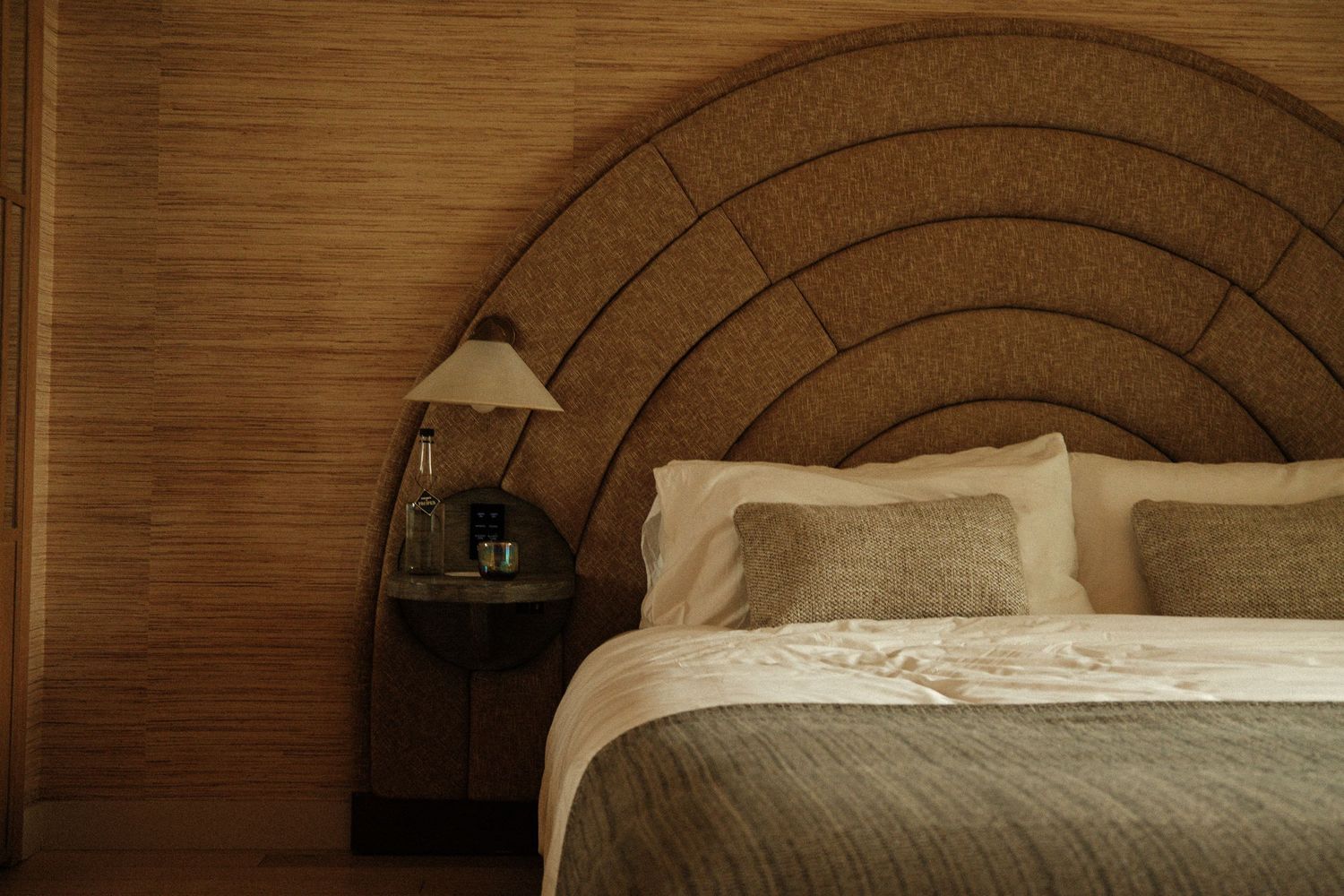 Luxury hotel bedroom with arched rattan headboard, neutral linen bedding, and warm bamboo wall paneling with bedside lamp.