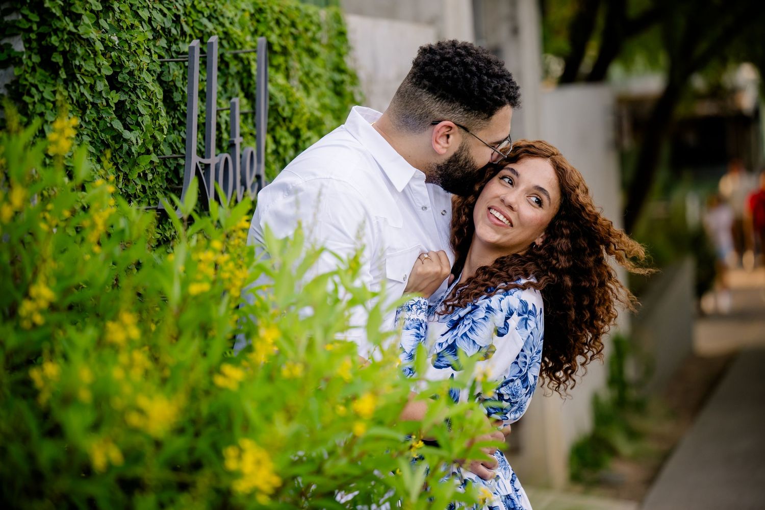 Couple sharing a romantic moment outdoors, surrounded by lush green foliage and yellow flowers on a sunny day.