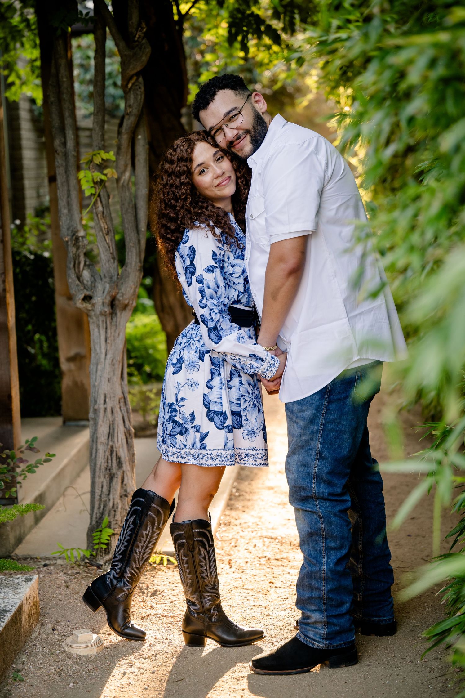 Couple embracing outdoors, woman in blue floral dress and cowboy boots, man in white shirt and jeans, surrounded by trees.
