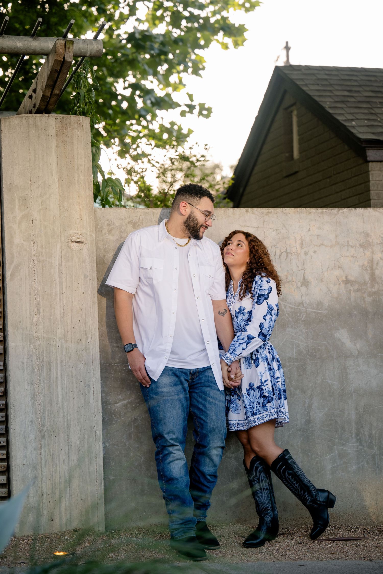 Couple holding hands and gazing at each other against a concrete wall, woman in floral dress and cowboy boots, man in white shirt.