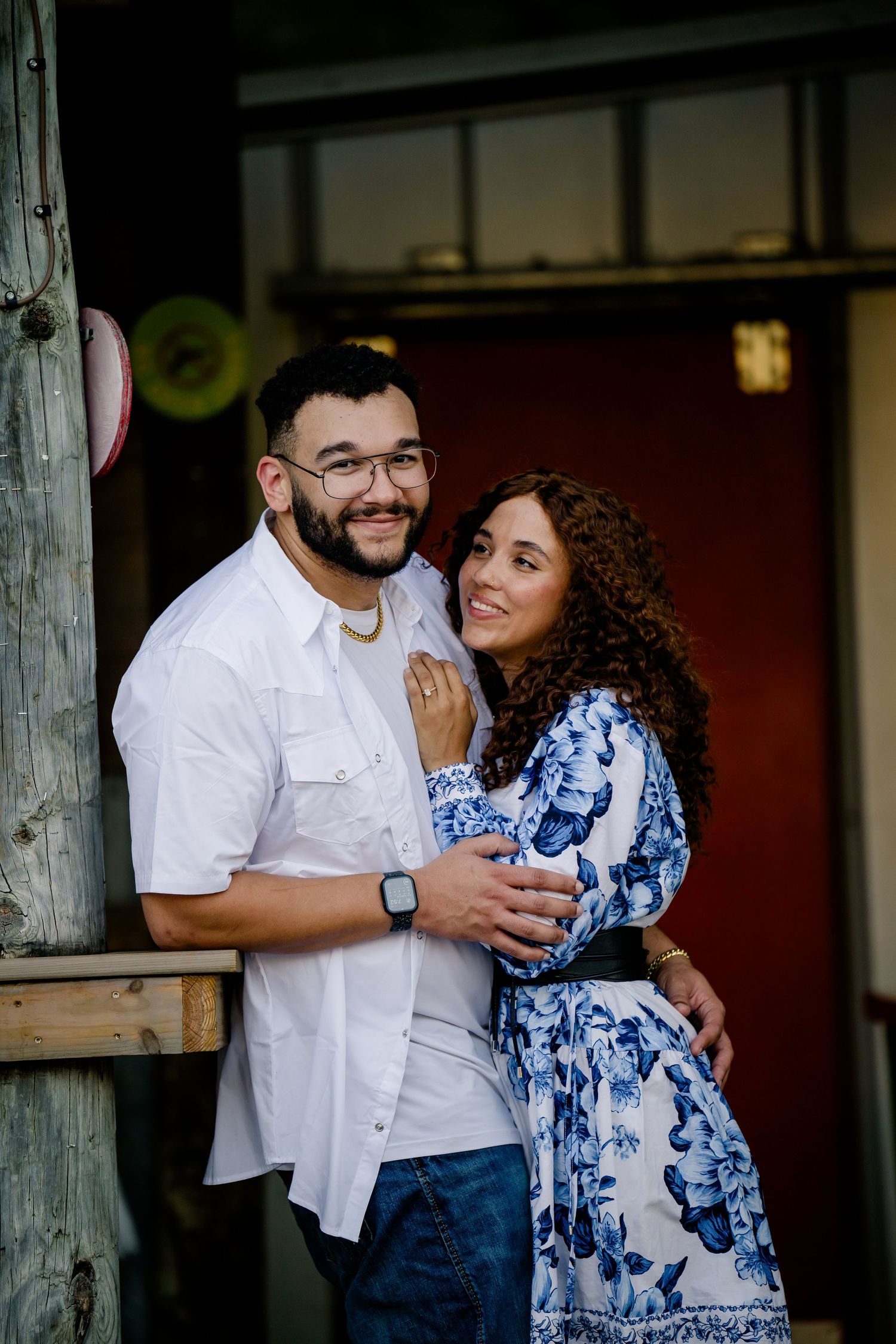 Smiling couple posing together on a porch, man in white shirt and woman in blue floral dress embracing warmly.