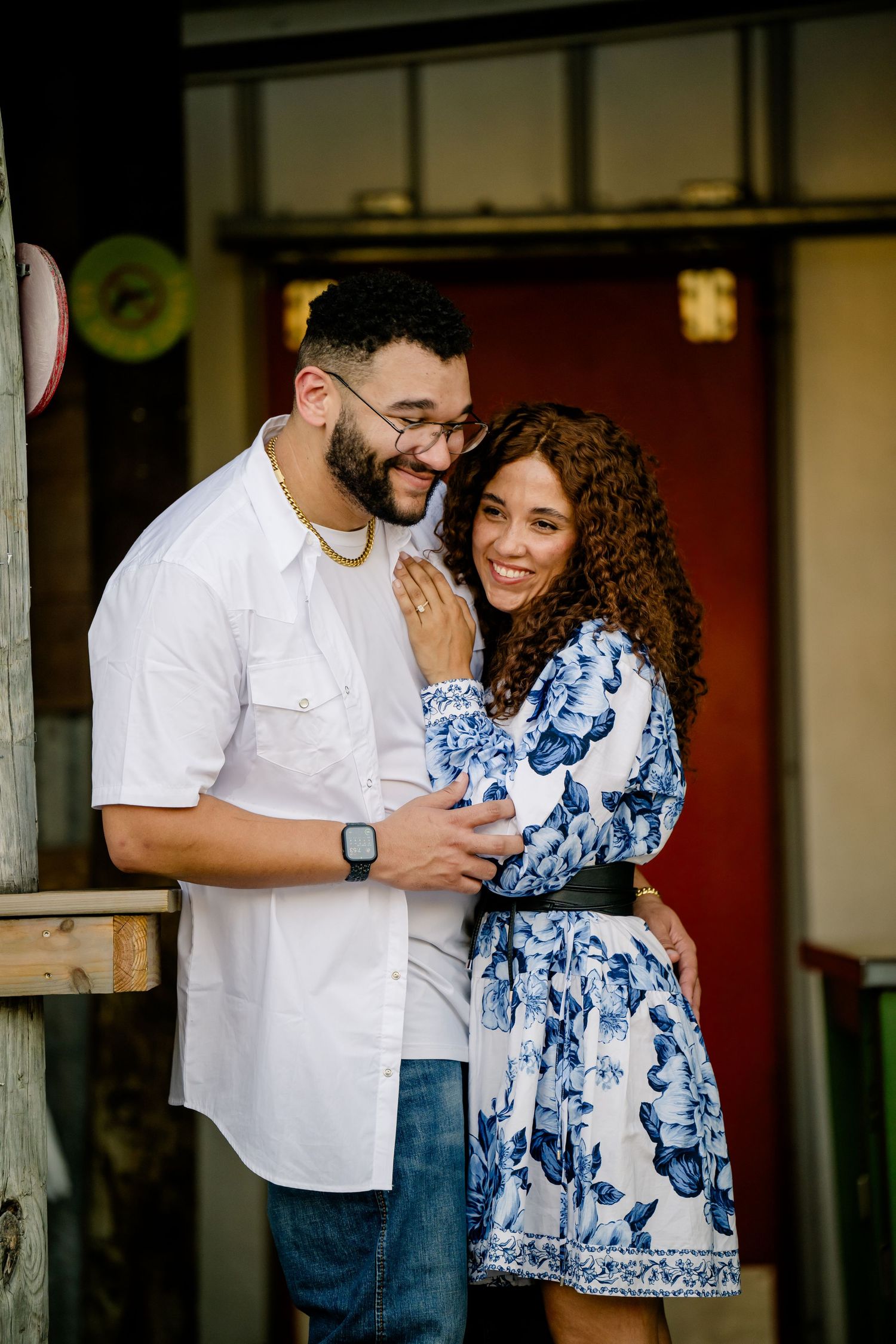 A couple smiles and embraces outdoors, man in white shirt and jeans, woman in blue floral dress, near a red door.