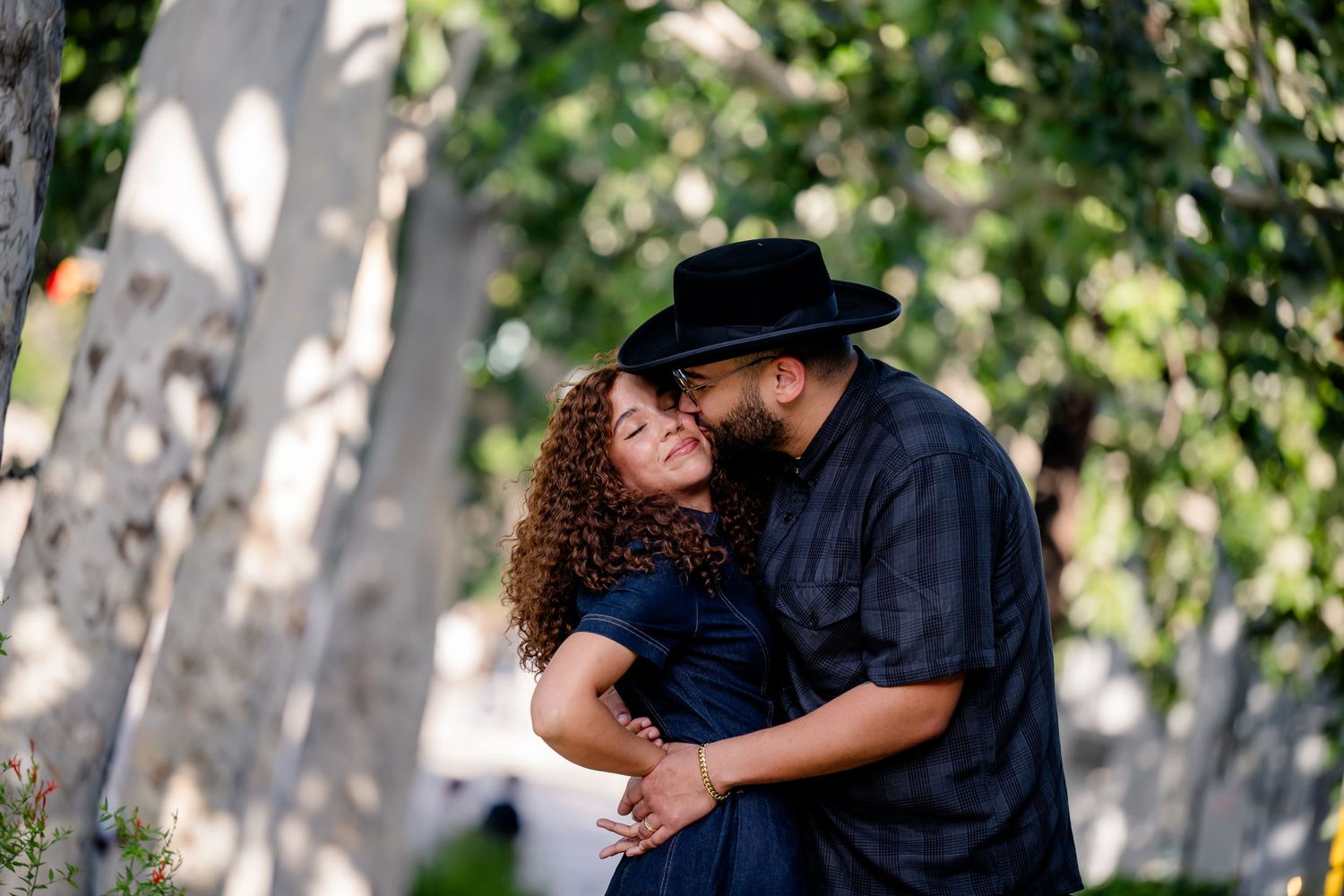 Couple sharing a romantic moment outdoors, woman with curly hair in navy dress, man in black hat and shirt, among trees.