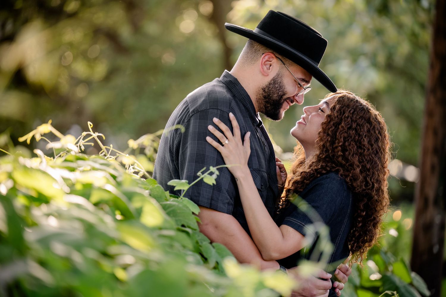A couple shares a romantic moment surrounded by lush green foliage, the man wearing a black hat gazing lovingly at the woman.
