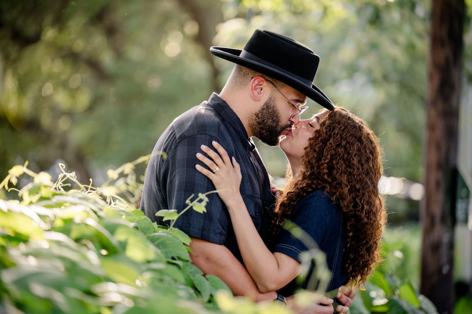A couple shares a romantic kiss outdoors, surrounded by lush green foliage, man wearing a black cowboy hat.