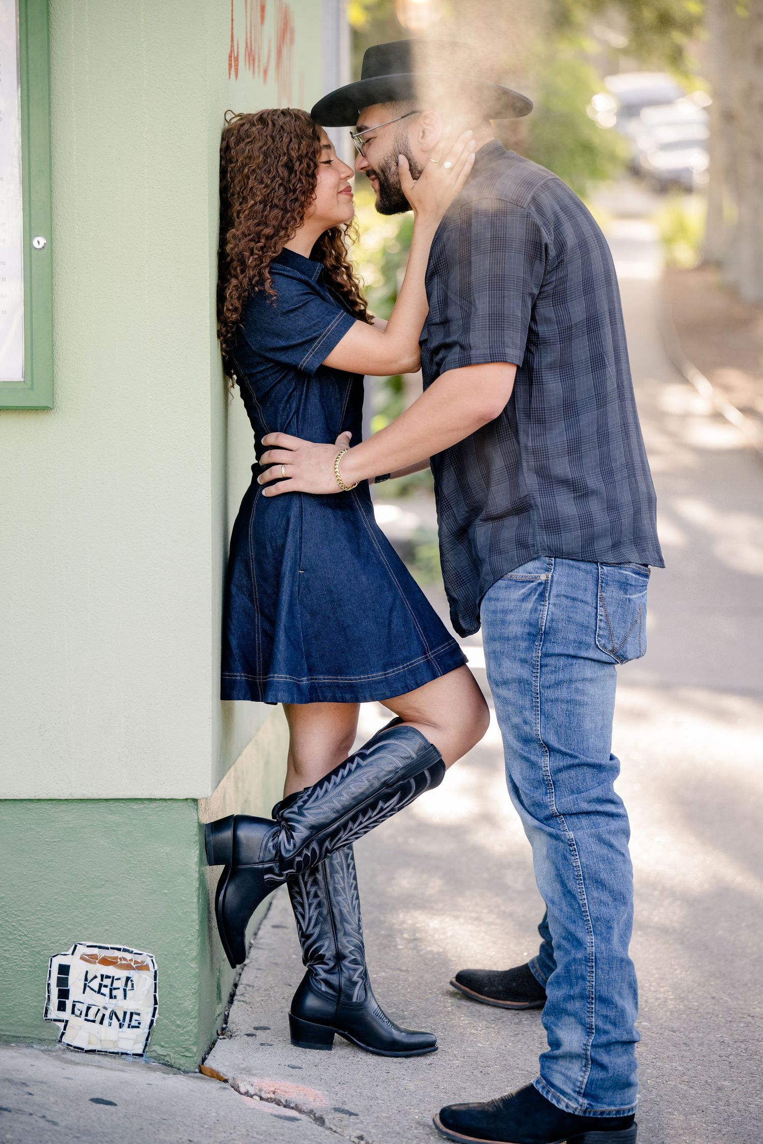Couple in western style sharing a romantic moment against a green building corner on a sunny sidewalk.