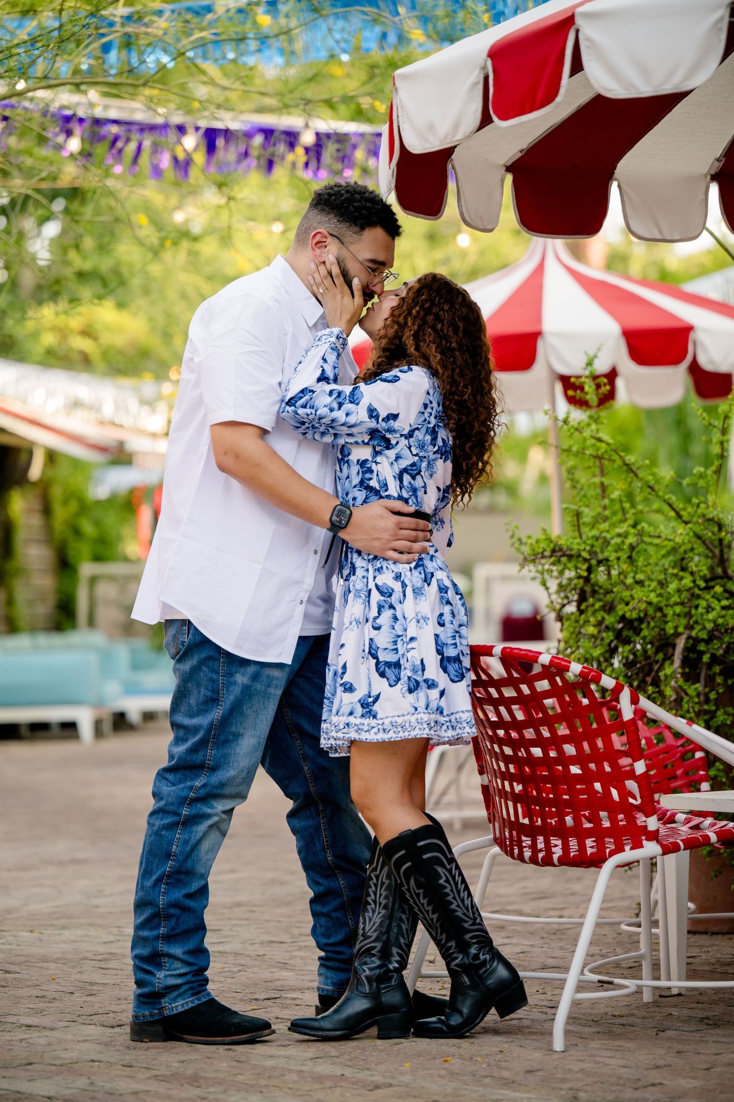 Couple sharing a romantic kiss outdoors near red striped umbrellas, woman in blue floral dress and cowboy boots.