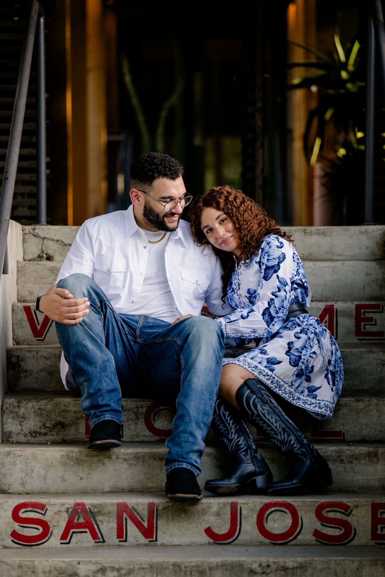Couple sitting closely on San Jose steps, woman in blue floral dress leaning on man in white shirt and jeans.