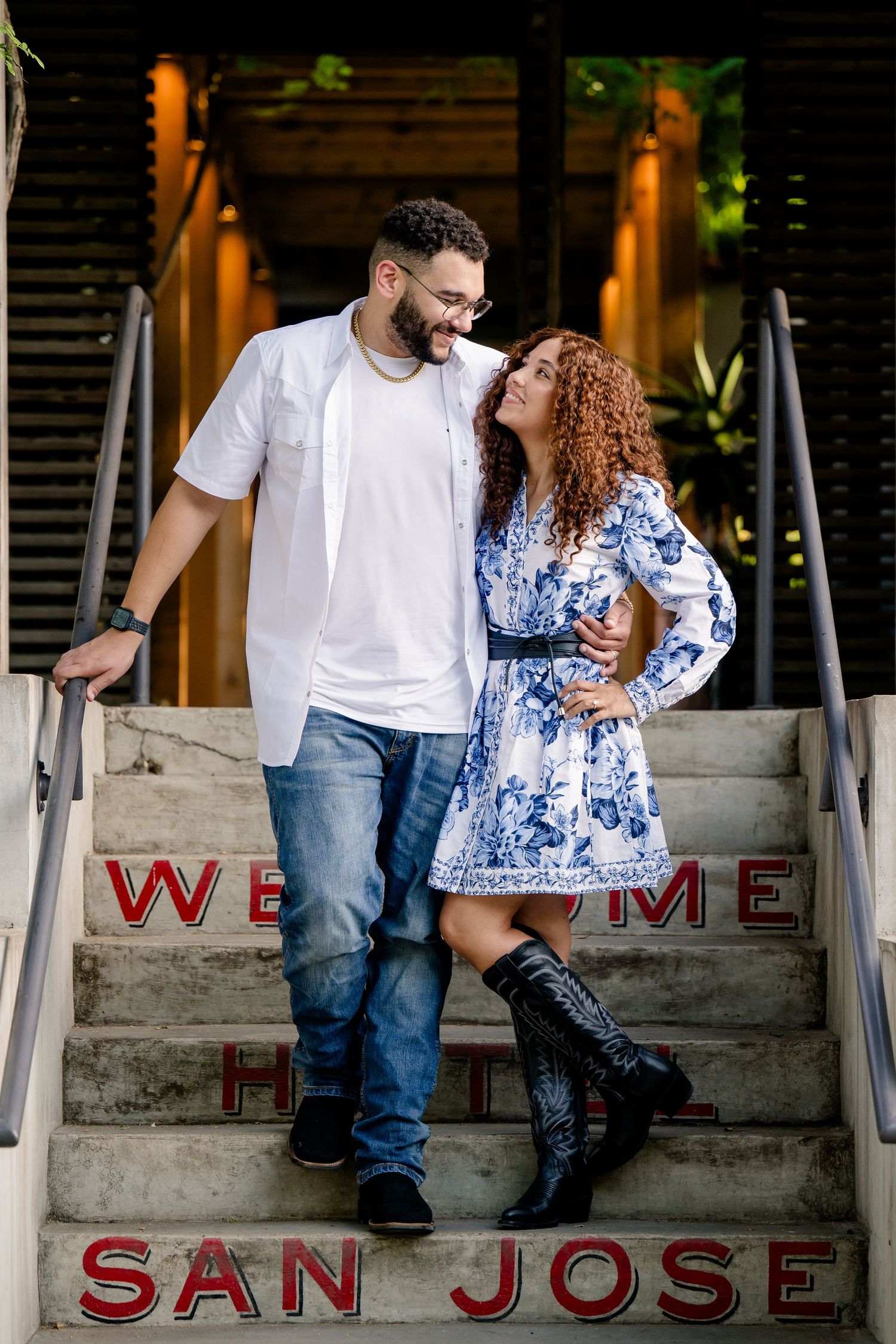 Couple smiling at each other on Welcome to Hotel San Jose steps, man in white shirt and woman in blue floral dress.