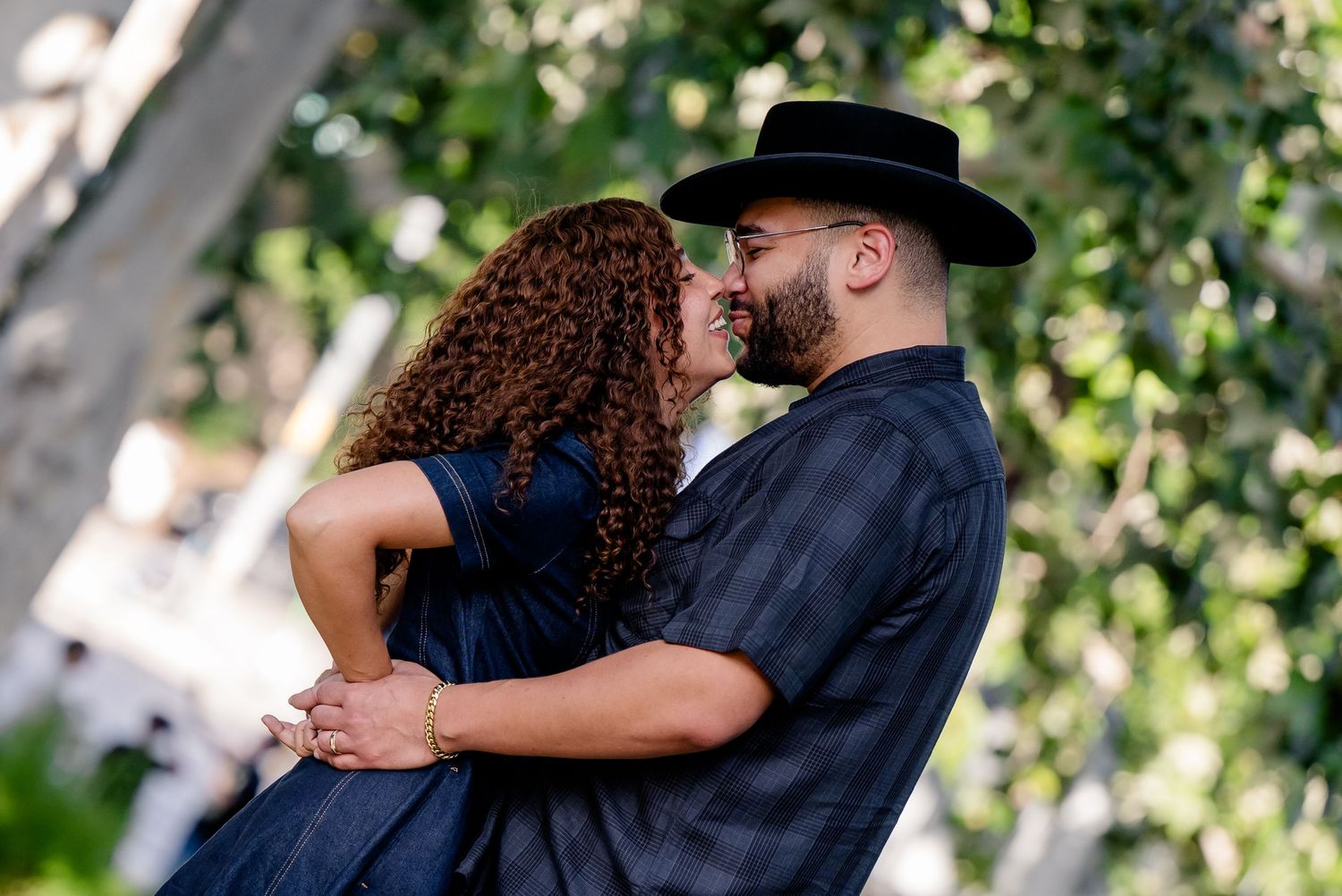 Couple sharing a romantic moment outdoors, man in black hat and shirt, woman in navy dress, surrounded by greenery.