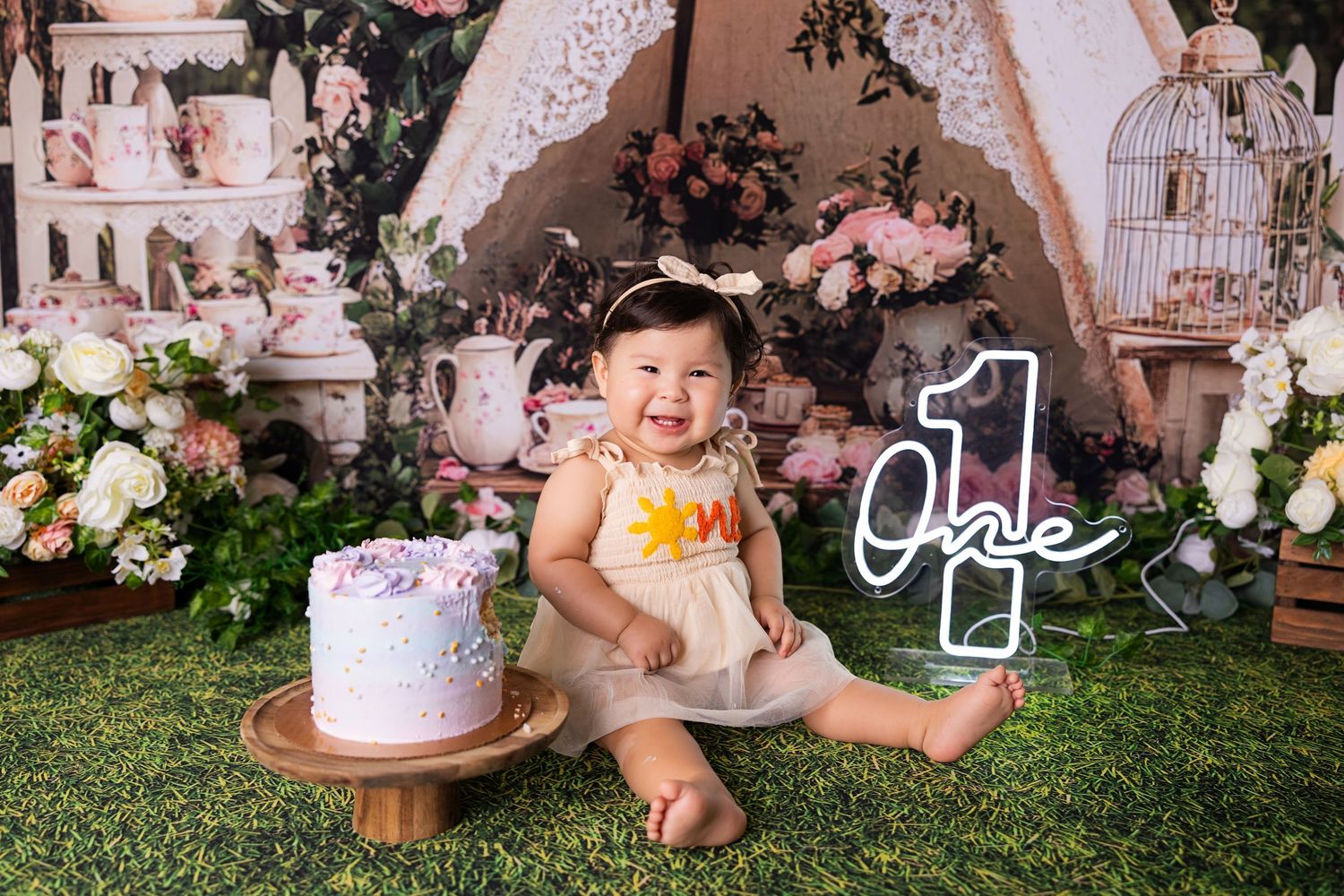 Baby girl in yellow dress smiles beside a purple smash cake at her first birthday celebration with floral decor.