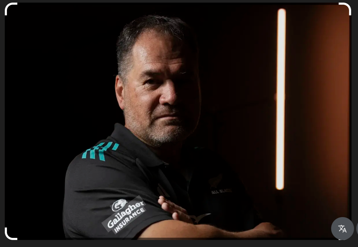 Man in All Blacks Gallagher Insurance rugby jersey poses with arms crossed against dark background.