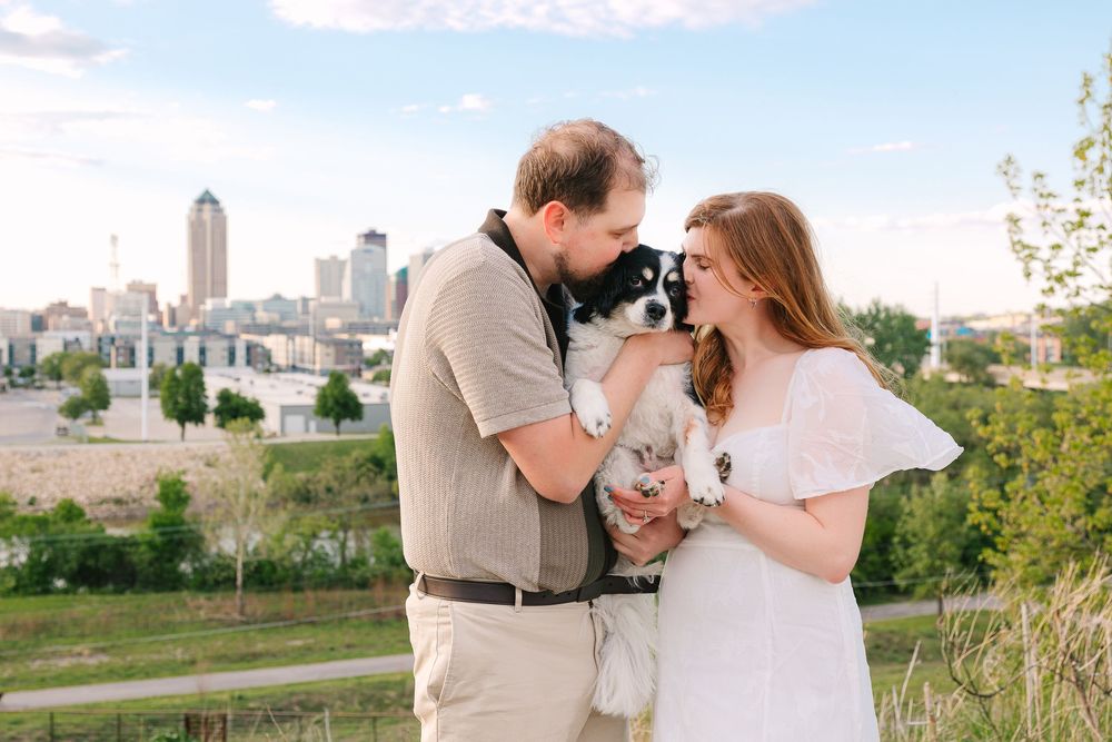 A couple hold and kiss their small black-and-white dog in front of the Des Moines skyline at MacRae Park.