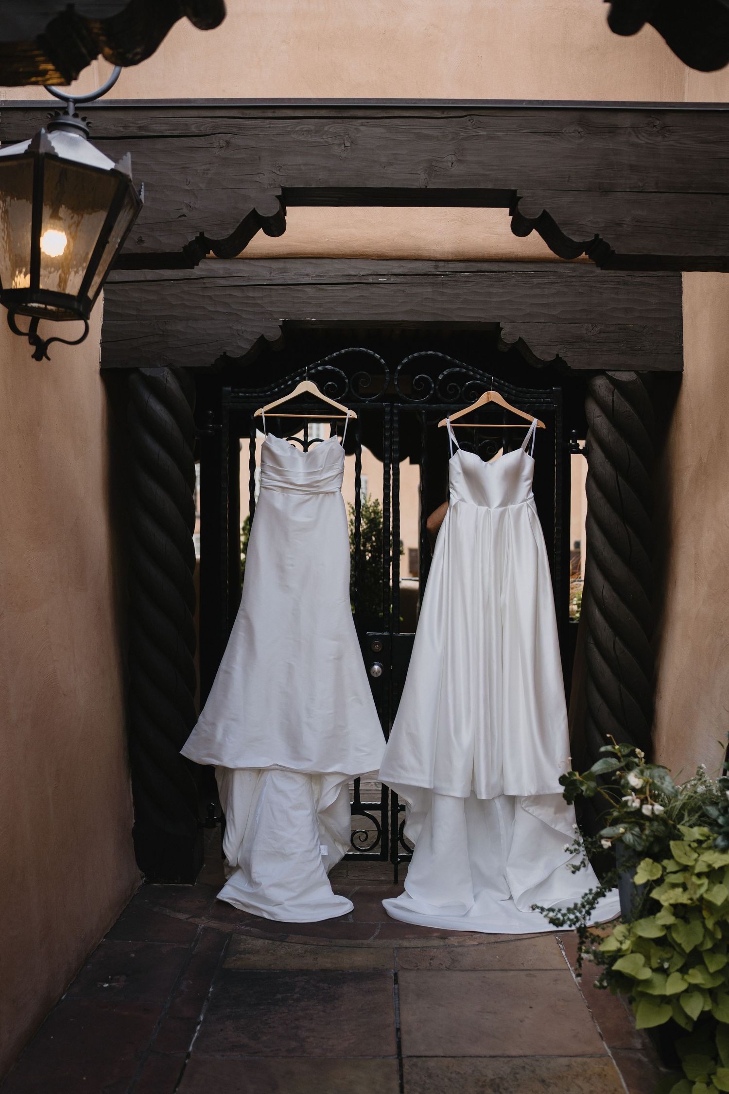 Two elegant white wedding gowns hanging on wooden hangers in a rustic doorway with wrought iron gates.