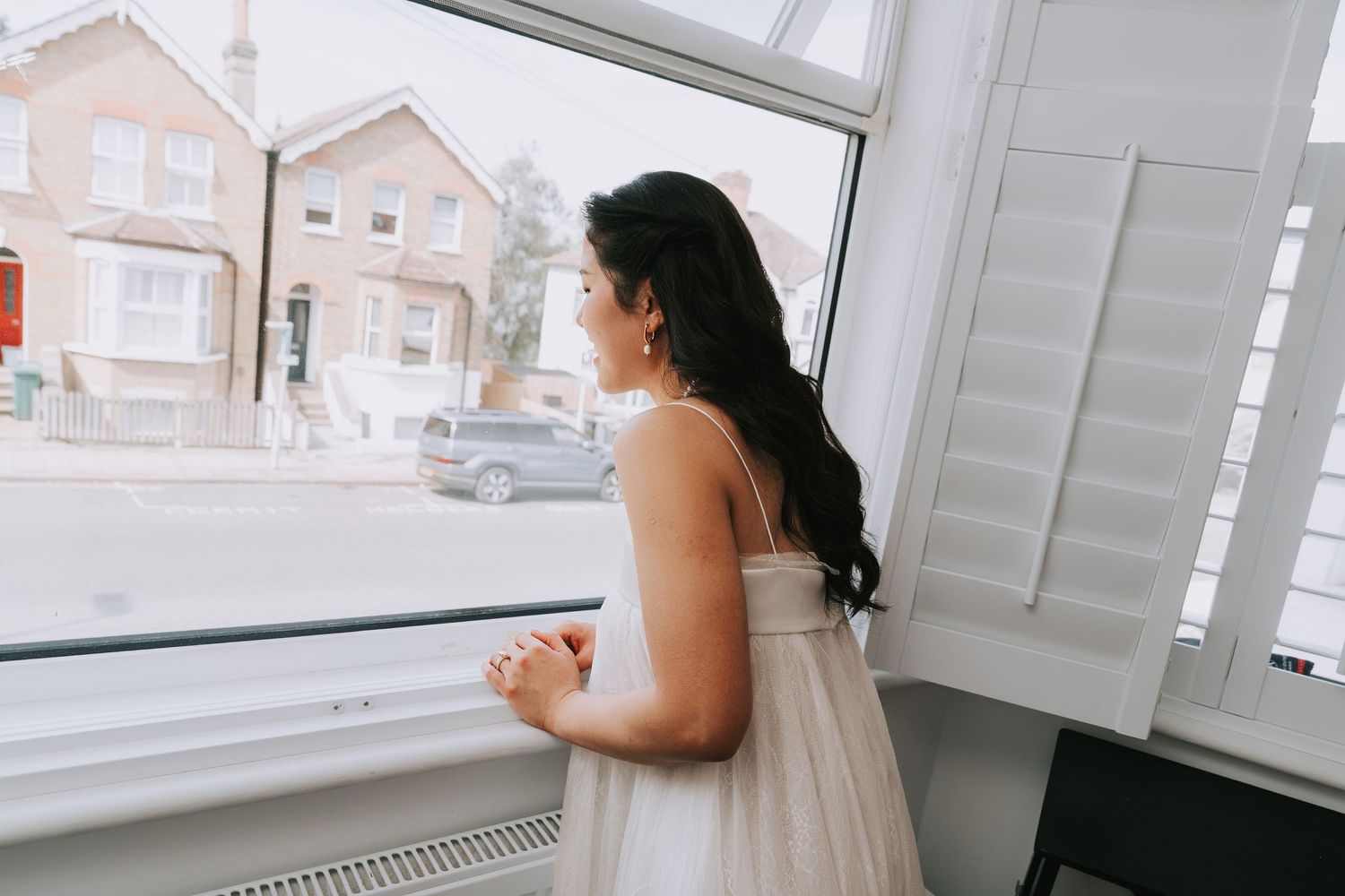Bride in white gown gazing out a large window on her wedding day.