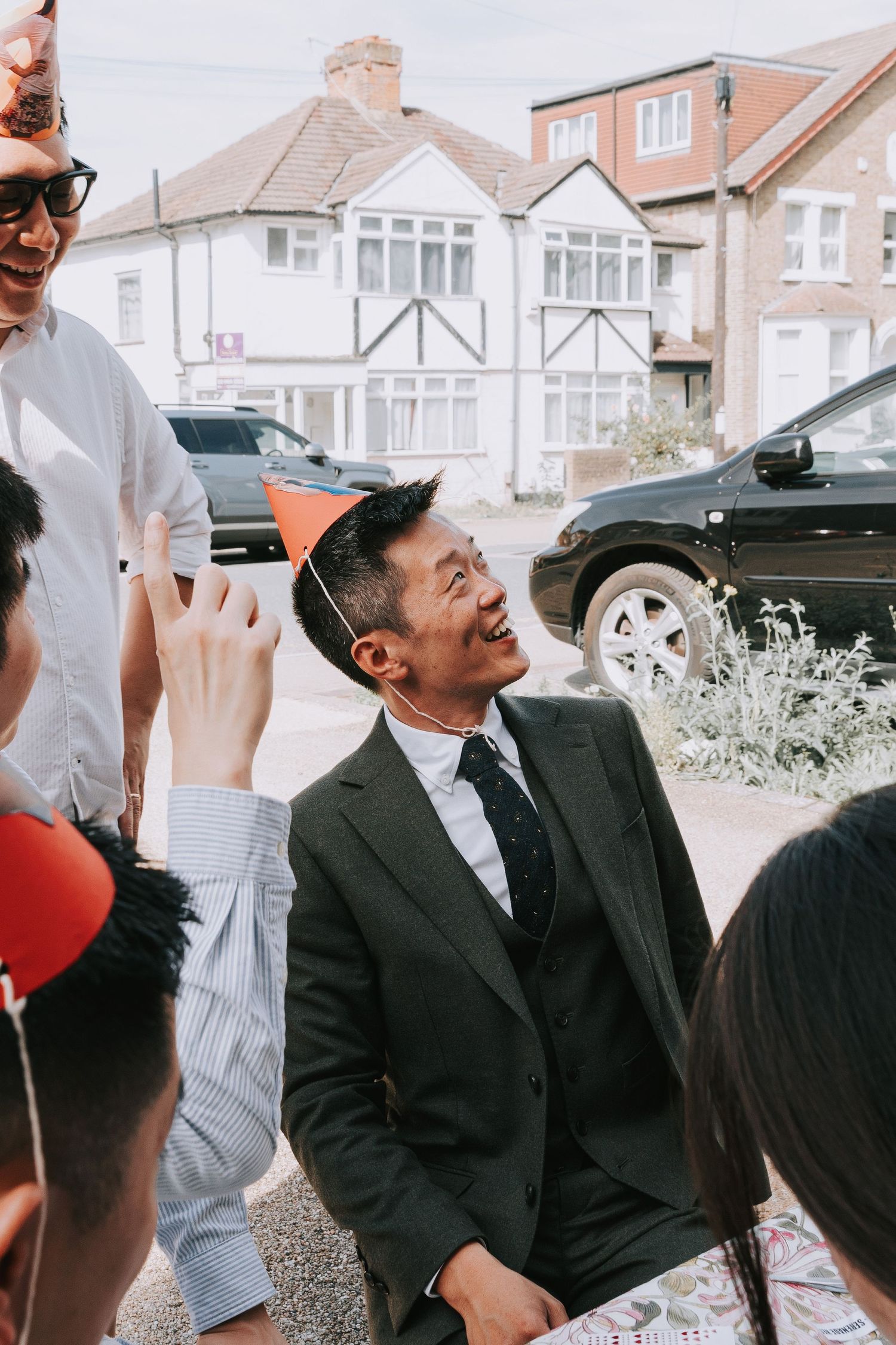 Smiling Asian man in dark suit and party hat celebrates outdoors with friends near suburban houses.