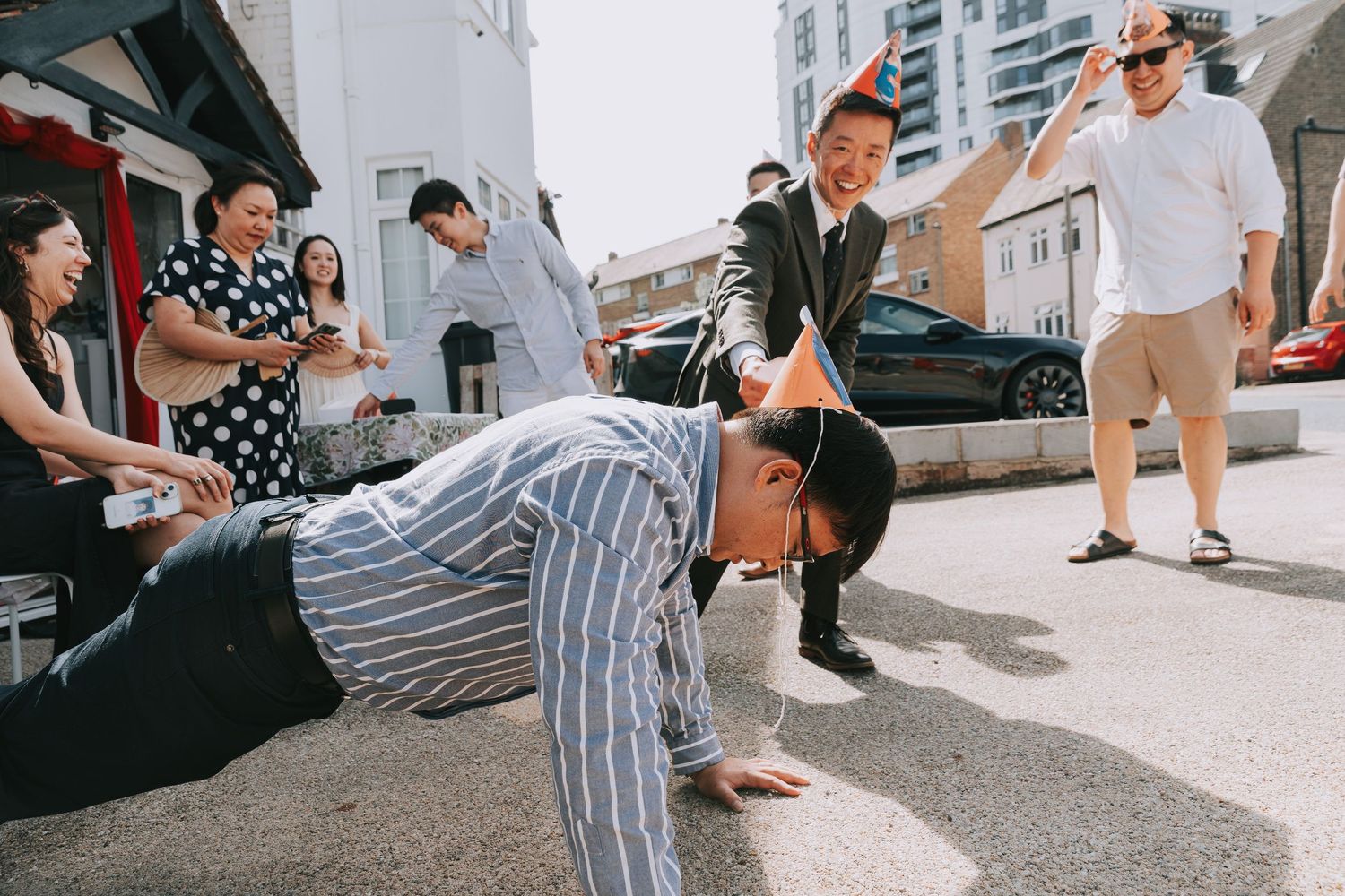 Man doing push-ups on street in striped shirt with party hat, surrounded by smiling onlookers at outdoor celebration.