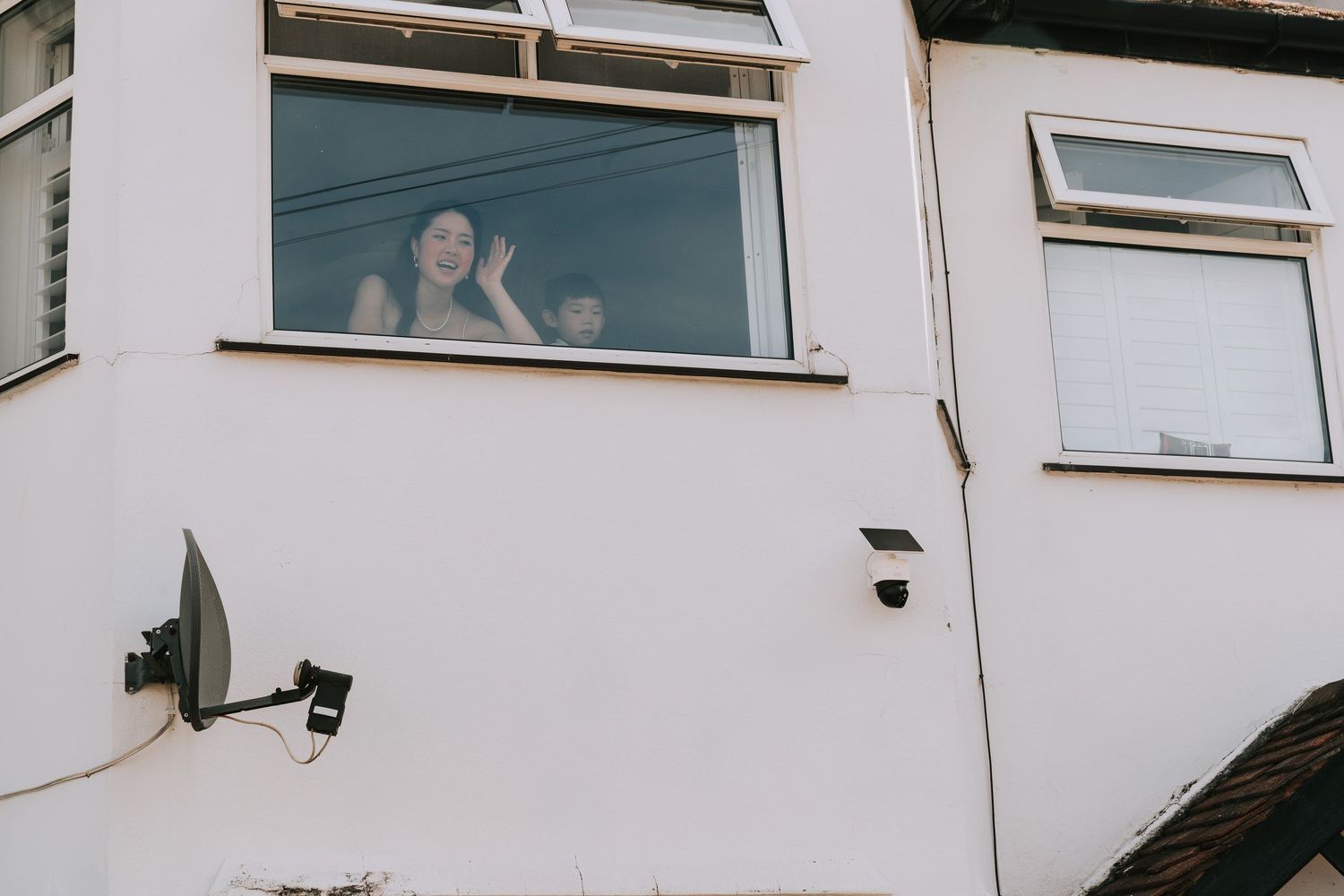 Woman waving from an upstairs window of a white residential building with a satellite dish and security camera.