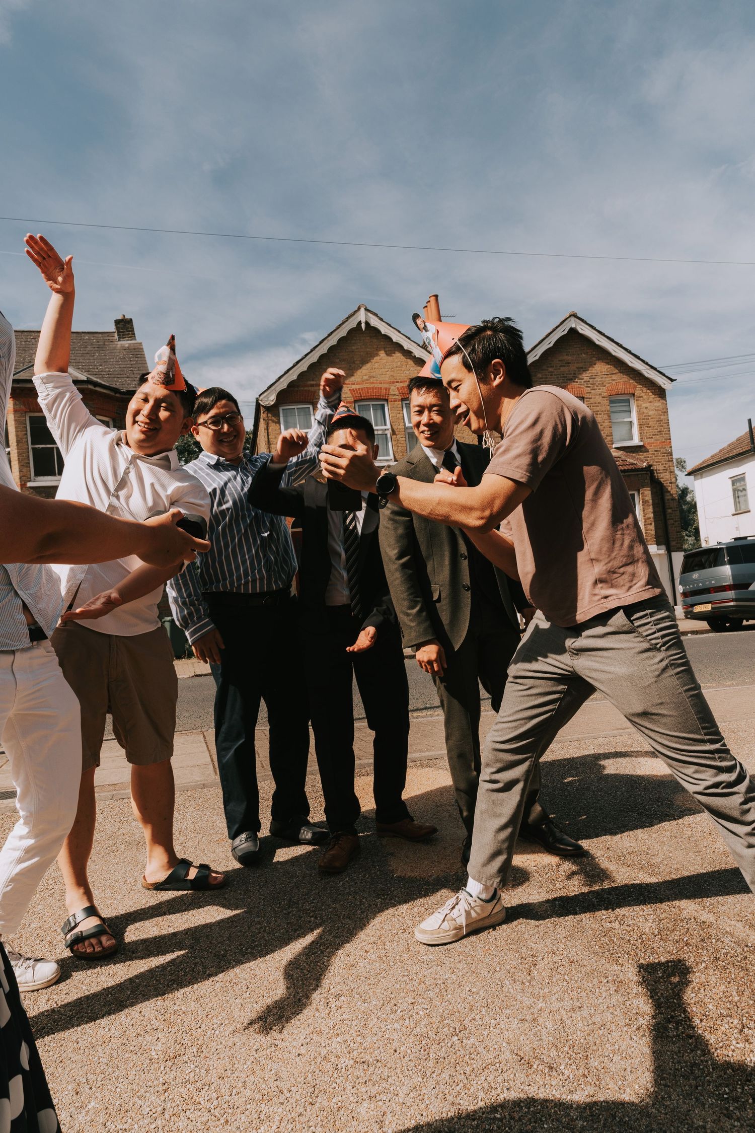 Group of young men celebrating outdoors on a street, laughing and dancing in front of brick houses on a sunny day.