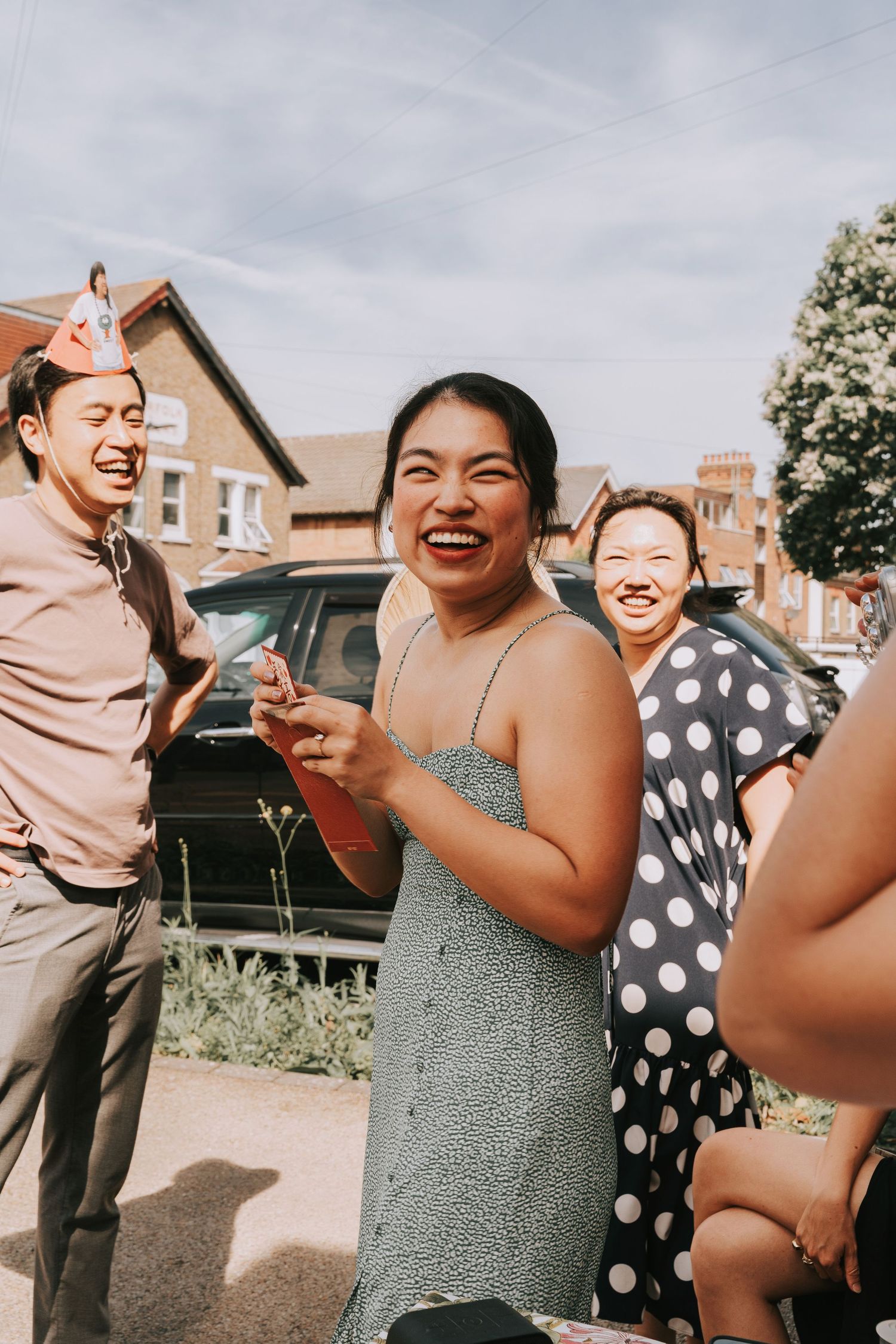 Group of joyful Asian friends laughing outdoors on a sunny day, woman in green dress holding red card.