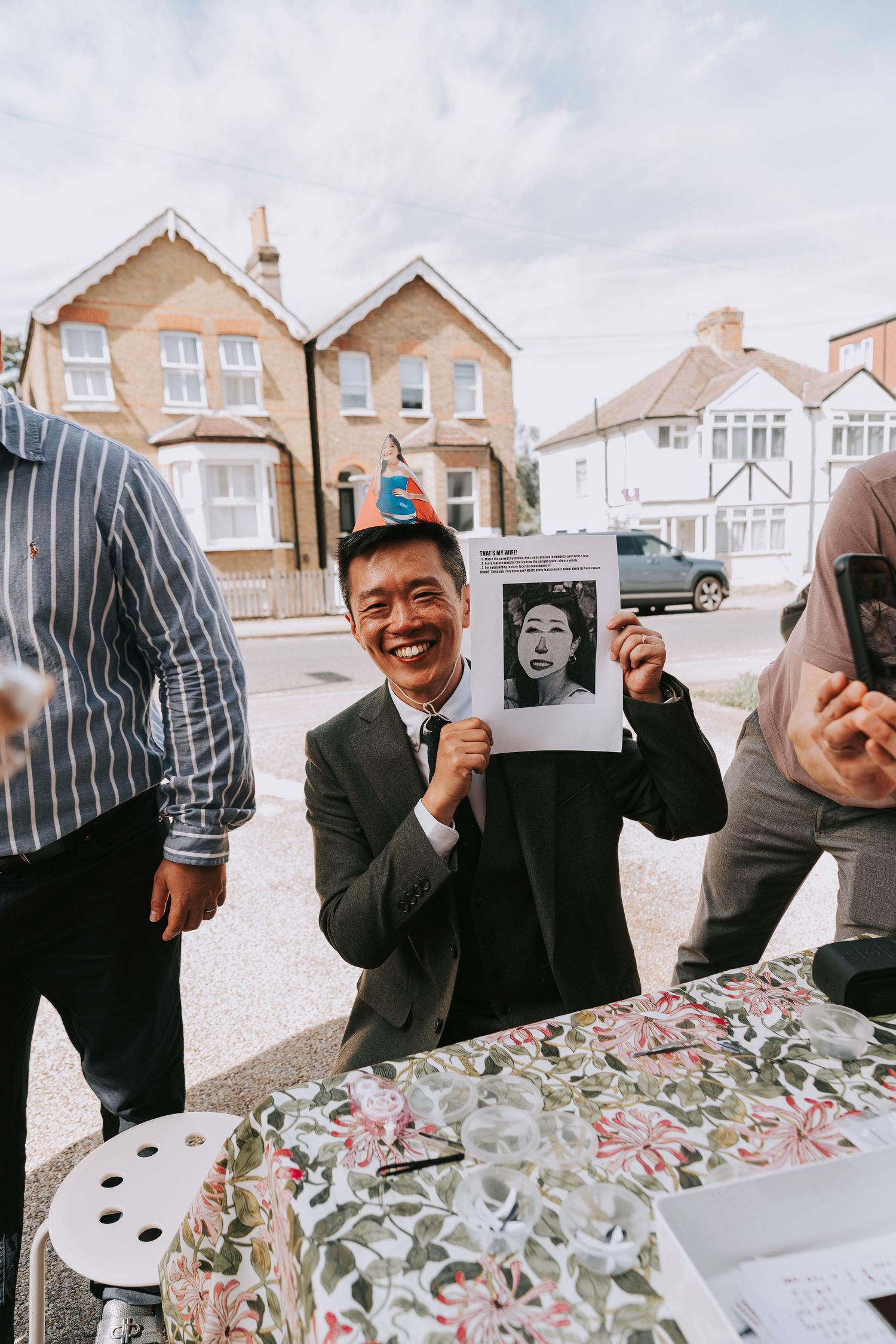 Smiling man in suit holding up a black and white photo at an outdoor table in a residential street.