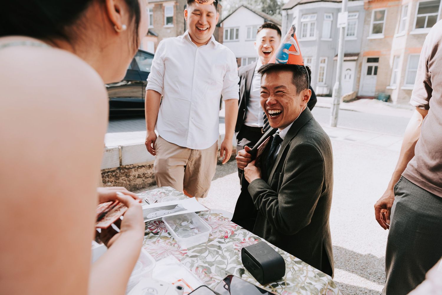 Man in birthday hat laughing joyfully at outdoor celebration with friends gathered around a table.