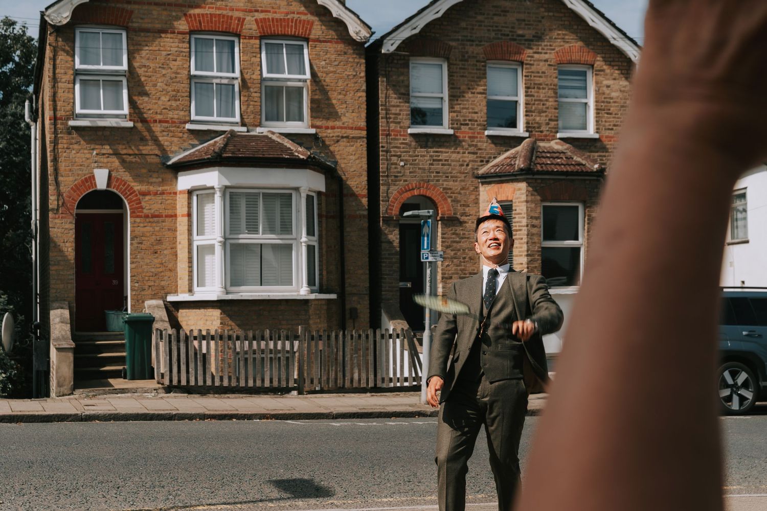 A well-dressed man in a suit stands on a sunny suburban street lined with classic brick townhouses.