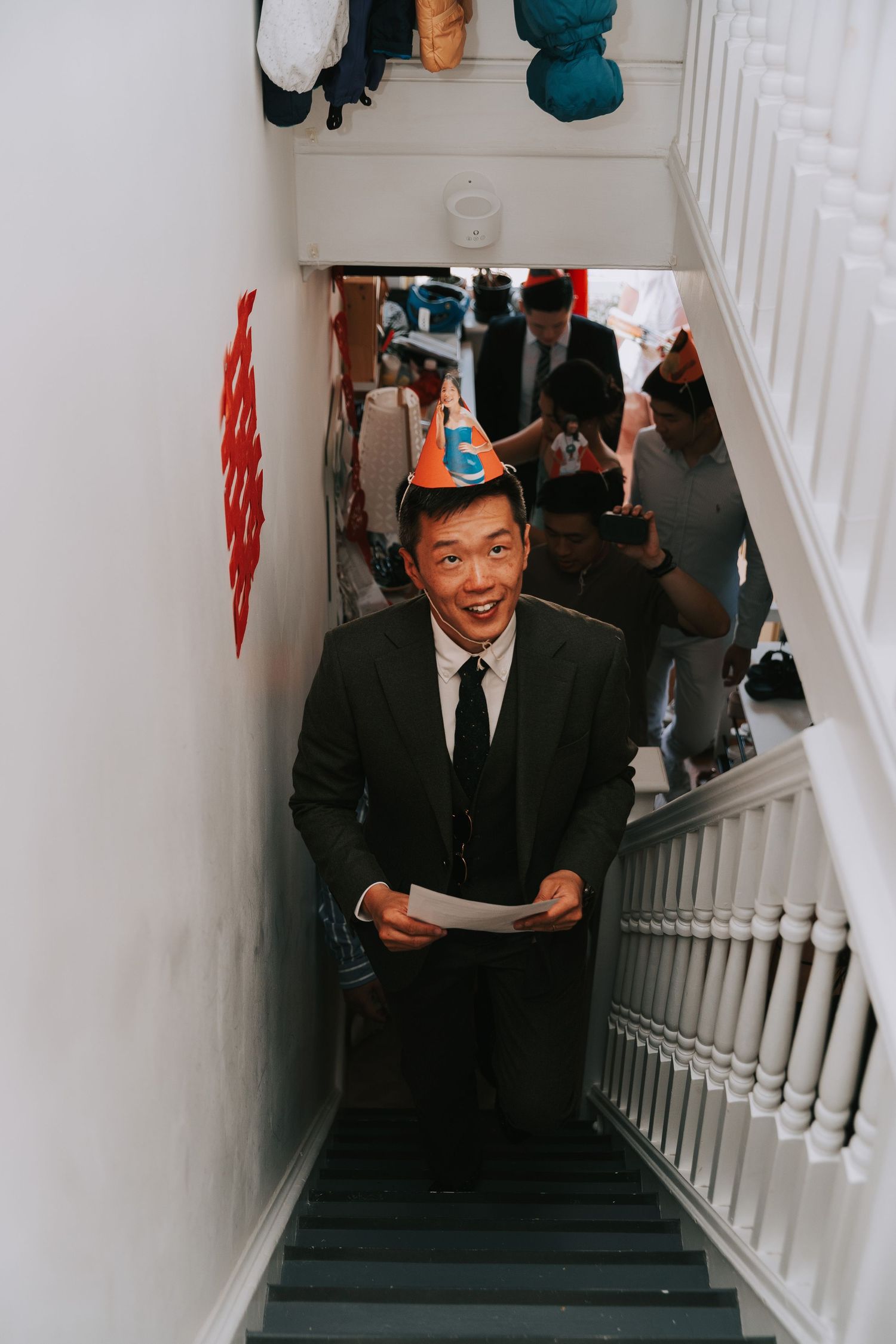 Man in suit and birthday hat smiling on staircase holding paper at a celebration.