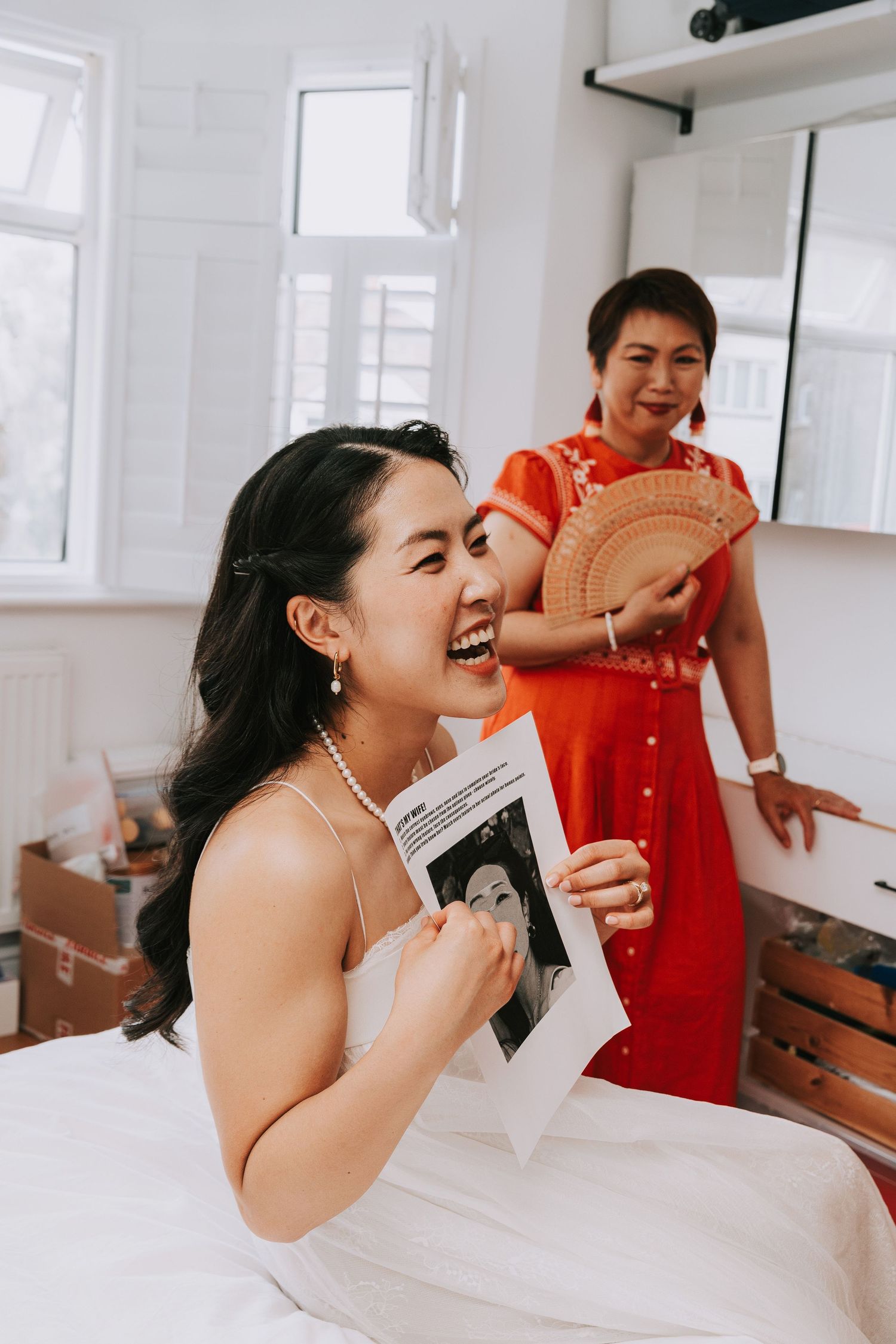 Bride in white dress laughing while holding photo, woman in red qipao behind her.