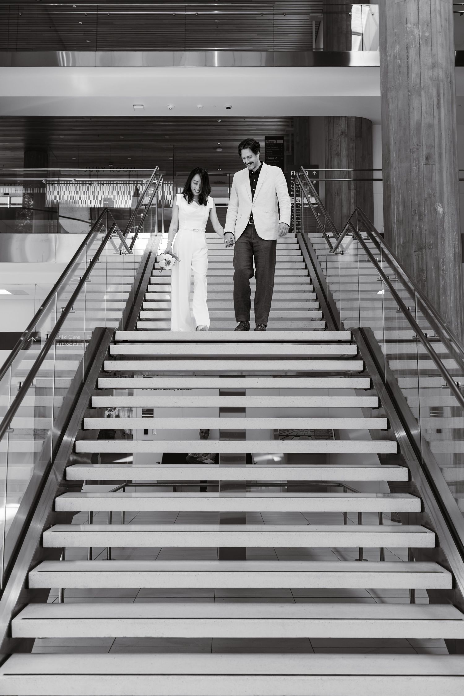 Black and white photo of a bride and groom holding hands while descending a grand staircase.