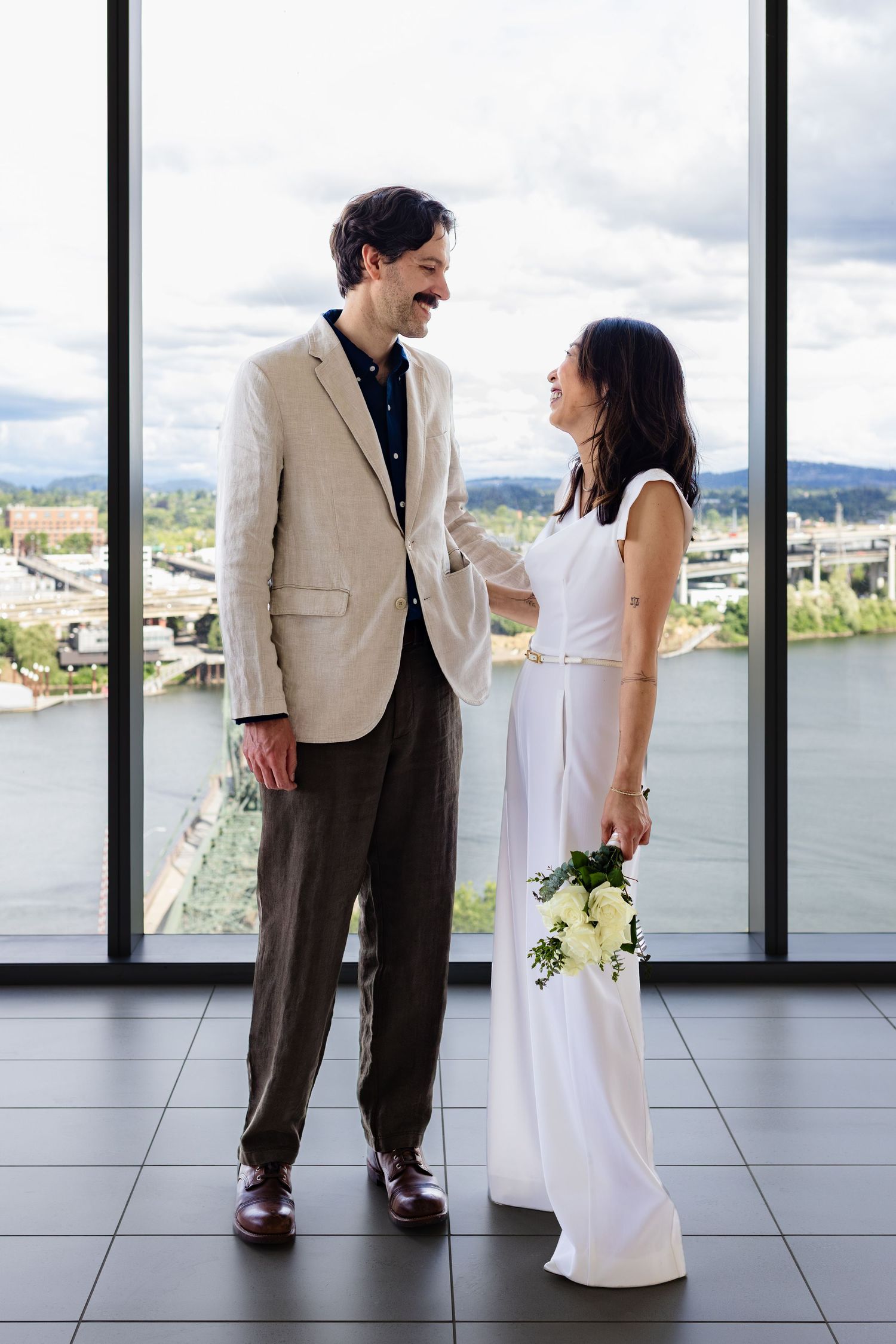 A smiling couple in wedding attire stands before a large window with a river view, bride holding white flowers.