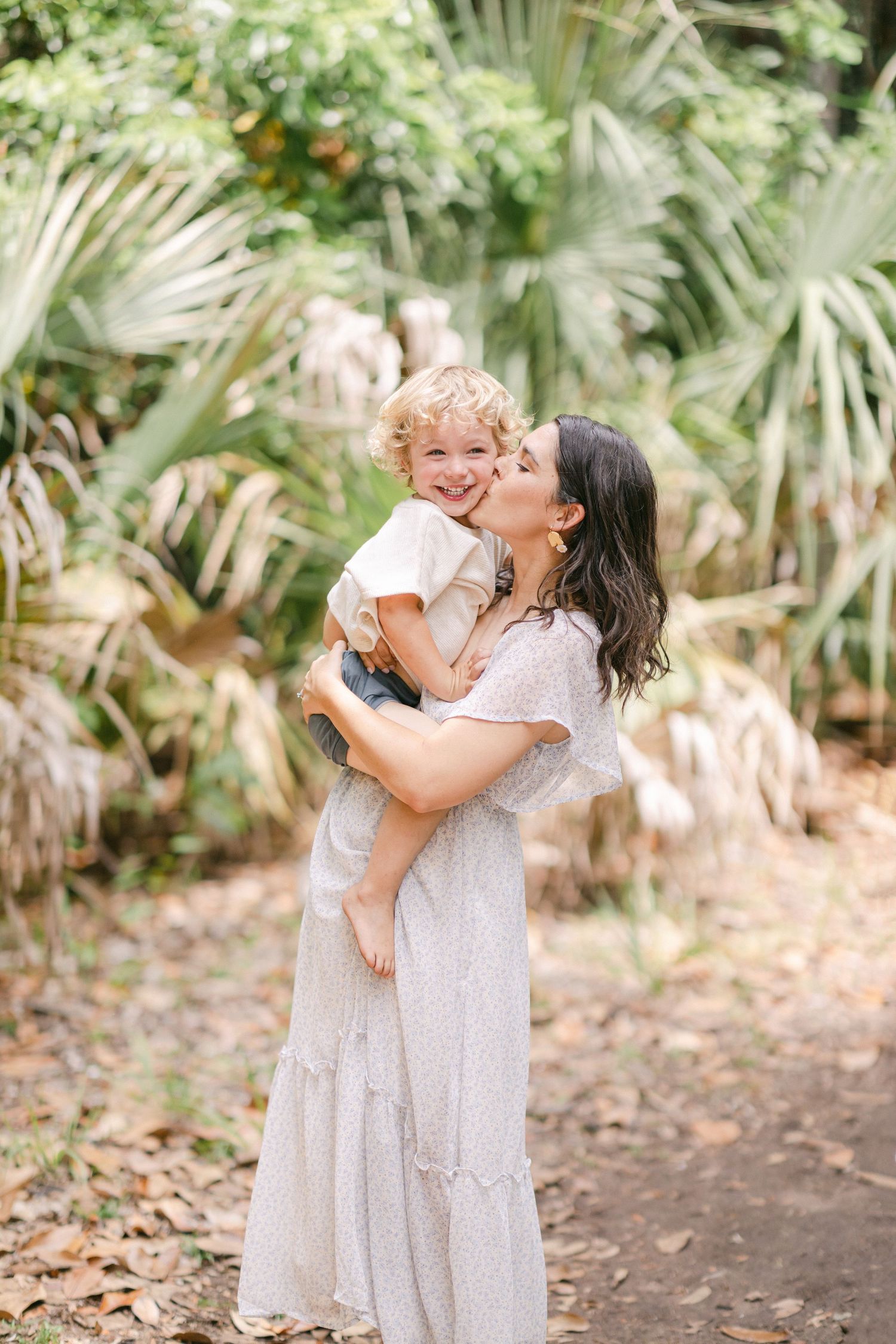 A dark-haired woman in a white dress kisses a laughing toddler boy amid lush tropical greenery.