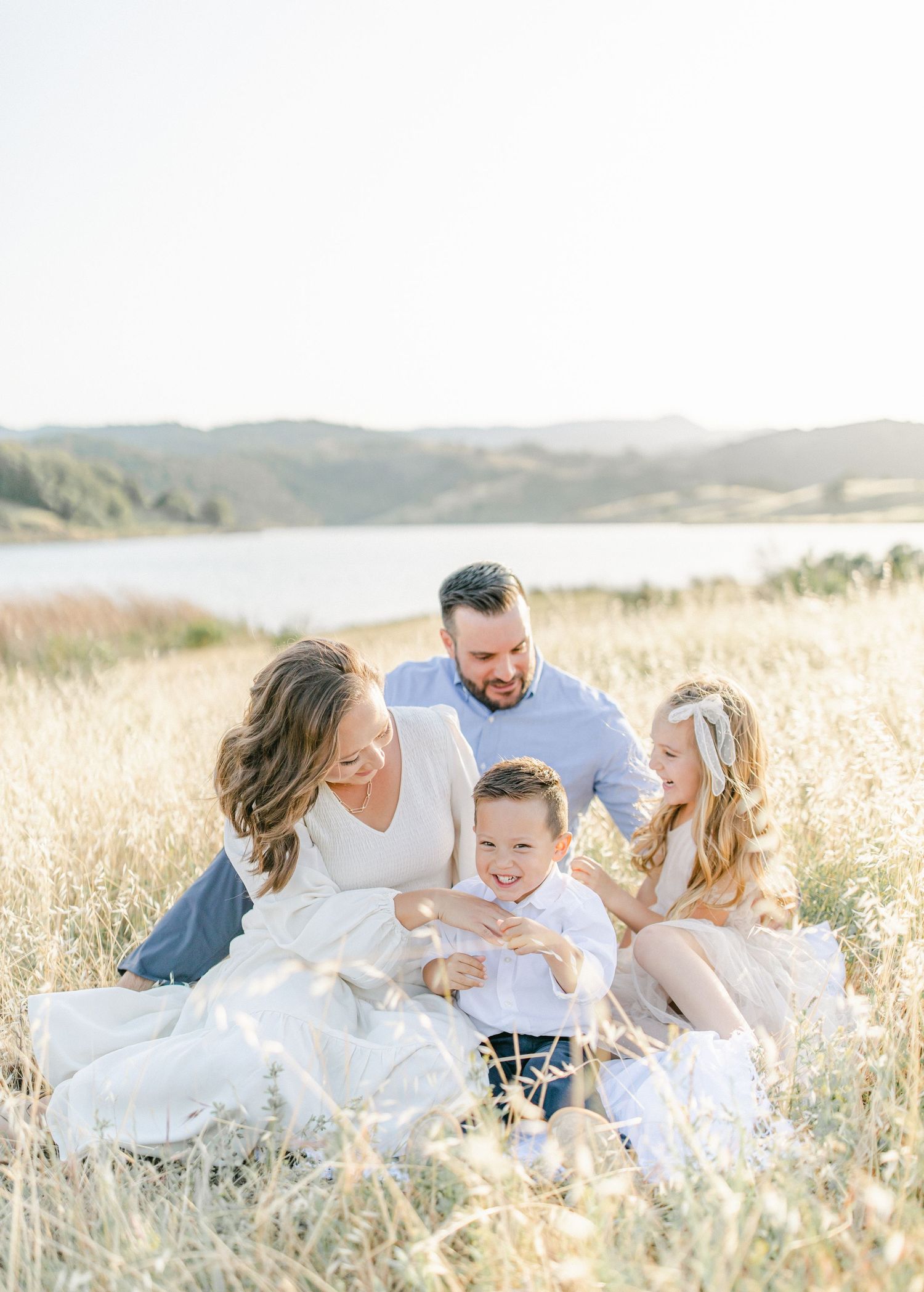 Family of four sitting in golden field laughing together, with scenic lake and rolling hills in background on sunny day.